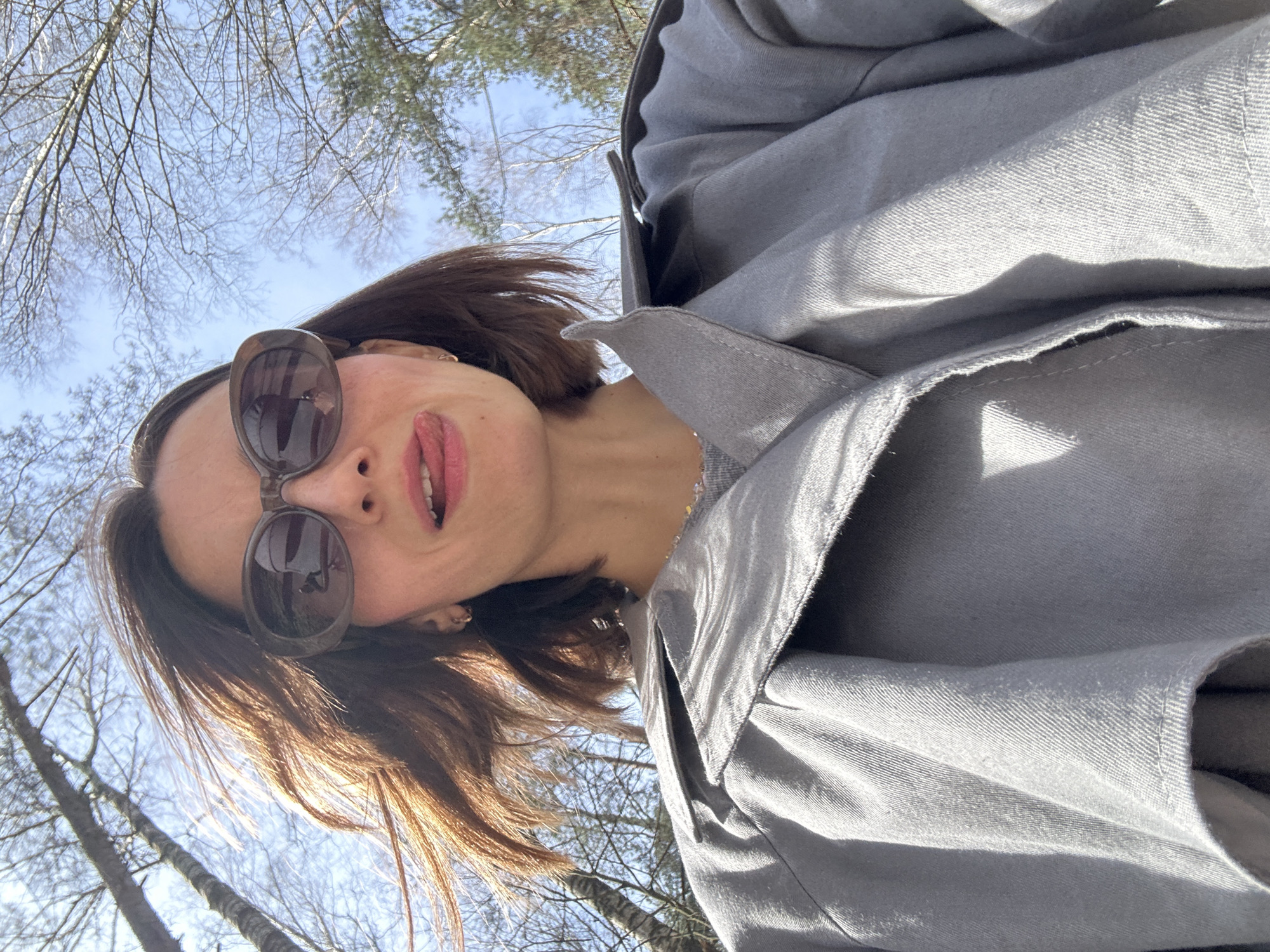The image presents a low-angle, close-up selfie of a person, primarily showing their head and upper torso. The subject's head is tilted sharply to their left (viewer's right), giving an unconventional perspective. They have medium-length brown hair and wear large, dark-framed sunglasses that cover their eyes. Their mouth is slightly open, revealing their tongue and teeth, and they have pink lipstick.
They are dressed in a light grey jacket or coat, made of a fabric with a visible twill weave, showing creases and folds. Bright sunlight casts distinct shadows and highlights on the garment.
The background consists of a clear, vibrant blue sky, indicative of a sunny day. Numerous bare tree branches, likely deciduous, are silhouetted against the sky, suggesting a late autumn, winter, or early spring season in an outdoor setting like a park or forest. The low photographic angle makes the sky and branches a prominent feature.
The activity is a casual outdoor selfie. The person's relaxed expression, with the slightly open mouth, conveys a playful or cheerful mood. The clear sky and strong sunlight confirm daytime conditions. Given the location context of Moscow, this scene portrays a sunny day in a natural area during the colder months when trees are leafless. There is no visible text in the image.