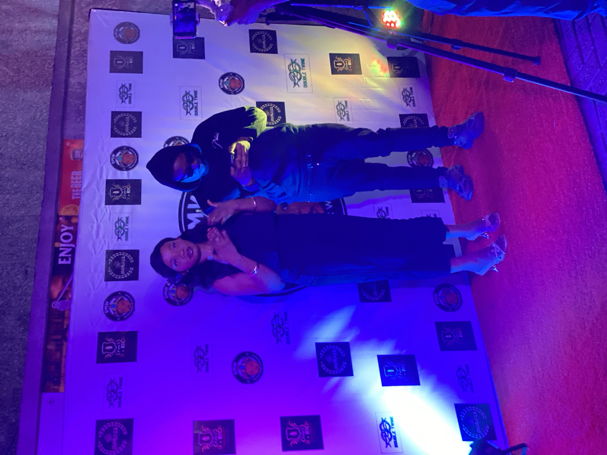 The image shows two people posing on a white backdrop decorated with logos, possibly for an event or promotion. A man in a dark hoodie and trousers is kneeling next to a woman in a dark outfit and heels. They are both looking towards the camera. A bright light is shining on the backdrop and the surrounding area is lit with a red or pink glow. The location is Iranla, Nigeria, based on the provided OCR which includes a reference to DMW, a Nigerian record label. Additionally, there is a partially visible sign with the word "ENJOY THE BEER". There are camera equipment visible in the background.