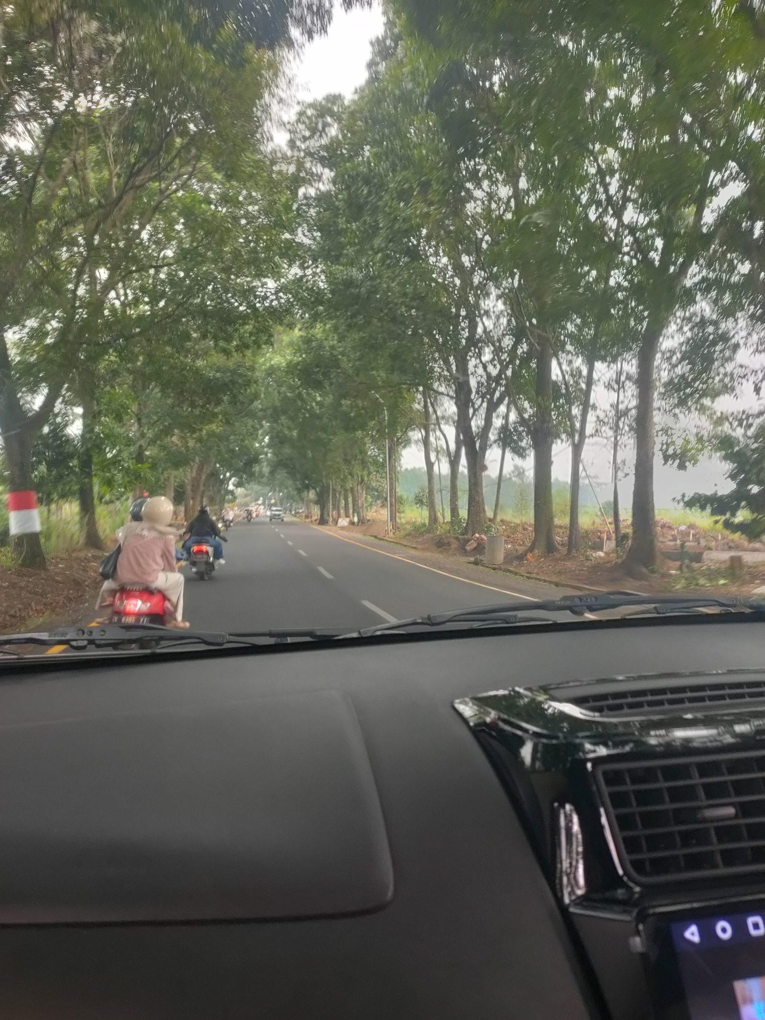 The image depicts a view from inside a car traveling on a road lined with trees in Subang, Indonesia. The road is paved with asphalt and marked with white lines. Several motorcycles are visible on the road, with two in the foreground. The first motorcycle, a red scooter, carries a person wearing a pink shirt and white pants, with a beige helmet. The second motorcycle, a dark-colored scooter, is ridden by a person in a black jacket and blue jeans. Further down the road, more motorcycles and a car are traveling. The environment is characterized by lush green trees on both sides of the road, with some rural scenery and construction debris visible on the right. The sky is overcast, suggesting a cloudy or hazy day. The dashboard of the car is visible in the foreground, including a portion of the air conditioning vent and a touchscreen display.