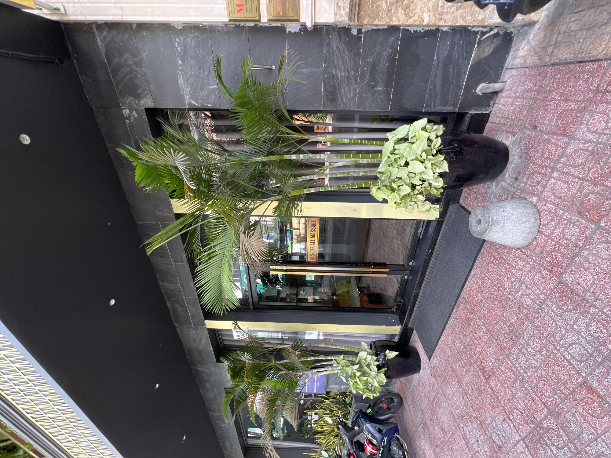 The image shows the entrance of a business in Nha Trang, Vietnam. A black facade with a decorative gold trim runs along the top. The entrance features glass double doors with gold trim. Several potted plants, including palm trees and leafy green plants, are positioned outside the doors. A dark mat is placed in front of the entrance. The ground is paved with red tiles featuring a geometric pattern. To the left of the entrance, two motorcycles are parked. A sign on the right door reads "10% OFF ALL MASSAGE". The overall scene depicts a commercial establishment with an inviting outdoor arrangement.