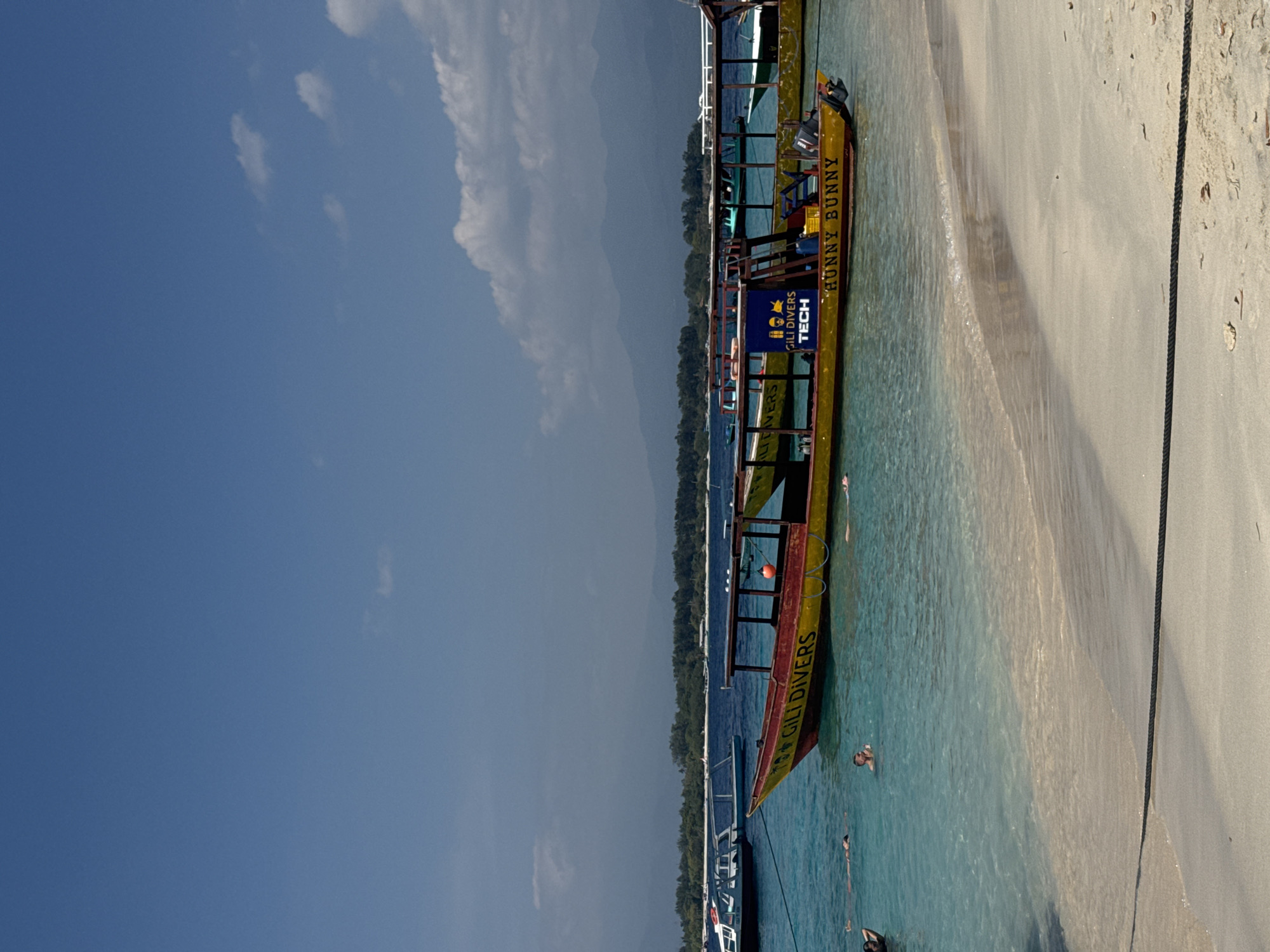 This image depicts a tropical beach scene in Gili Trawangan, Indonesia, under a clear blue sky with scattered clouds. In the foreground, a sandy beach meets the turquoise water. A dark rope stretches across the sand, likely a mooring line. The water is shallow and transparent near the shore, gradually deepening offshore.
Several people are visible swimming and relaxing in the water. In the middle ground, a yellow and red wooden boat is anchored, identified by the text "GILI DIVERS TECH" and "HUNNY BUNNY" on its side. This boat appears to be a dive or transport vessel. Another similar boat is visible further in the distance.
Behind the boats, a lush green coastline with trees is visible, and beyond that, distant mountains form the horizon. The overall atmosphere is serene and indicative of a popular tourist destination, likely a popular spot for diving and water activities. The sun is bright, casting reflections on the water and illuminating the scene.