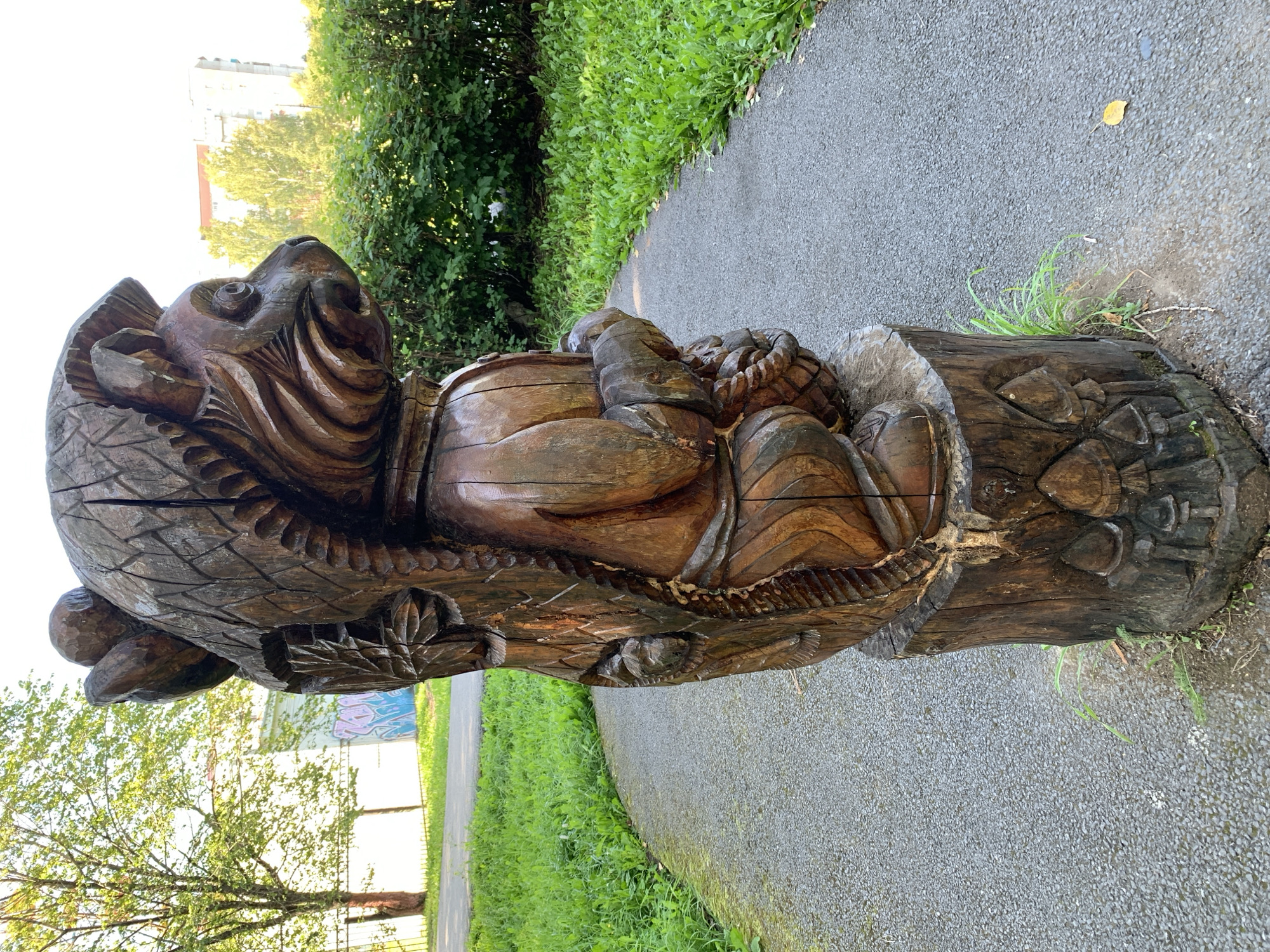 A carved wooden sculpture of a squirrel holding a nut stands on a tree stump along a paved pathway in a park. The sculpture is brown and weathered, with intricate details depicting the squirrel's fur, face, and paws. The squirrel is positioned in a crouched stance, its bushy tail curving upwards. The tree stump base is also carved with mushroom motifs. The pathway curves to the right and is flanked by green grass and lush bushes. In the background, a tall tree with green foliage stands to the left, and a modern building is visible behind the bushes to the right. The sky is bright and clear, suggesting a sunny day. This sculpture is located in the city of Prokopyevsk, Russia.