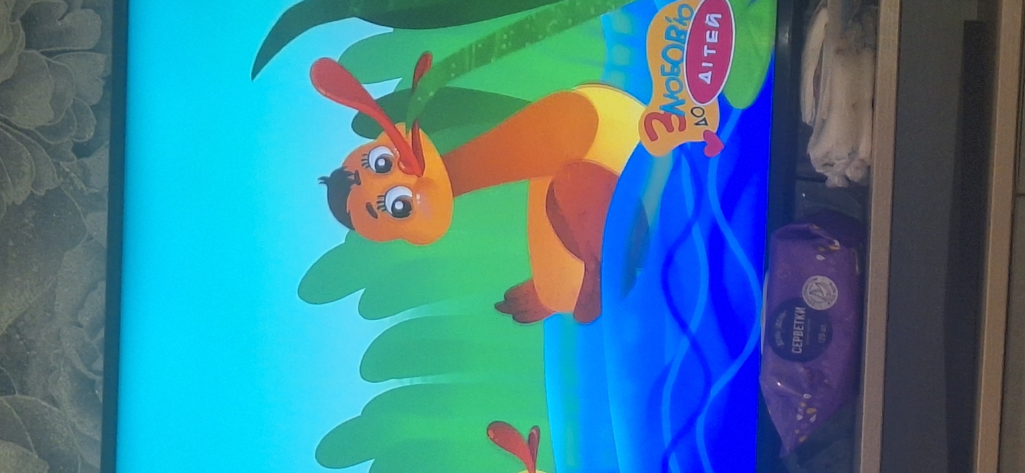 The image shows a cartoon animation of a duck swimming in blue water. The duck is orange and yellow with a black tuft of hair and large eyes. Behind the duck are green reeds. In the lower right corner of the screen is a logo that reads "З ЛЮБОВ'Ю ДО ДІТЕЙ" (With love for children). Below the screen, on a shelf, is a purple package of wet wipes with Russian text that includes "СЕРБЕТКИ" (wipes). To the right of the wipes is a bundle of white cloth items. The image is a photograph of a television screen displaying this content. There is no information in the image that suggests it is located in Bila Tserkva, Ukraine.