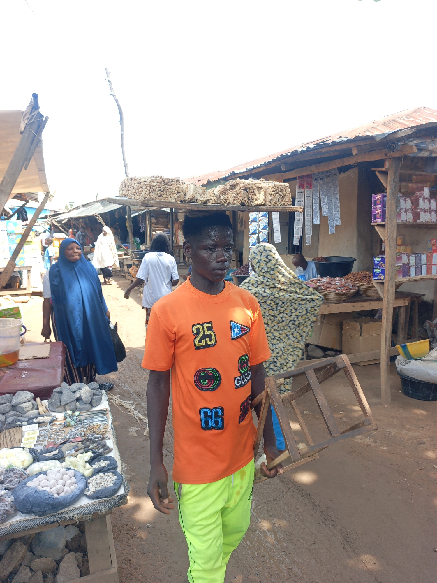 The image depicts a street scene in Akwanga, Nigeria. In the foreground, a young man wearing an orange t-shirt with the numbers 25 and 66 and green pants is carrying a wooden structure resembling a stool. Behind him, a woman in a patterned headscarf is visible. To the left, another woman in a blue garment walks alongside a table laden with various items for sale, including rocks, bracelets, and bags filled with white items resembling eggs or bulbs. In the background, stalls line the street, with one displaying bundles of sticks and another offering various goods like nuts, dried foods, and packaged items on shelves. Other people are visible in the background, moving through the market. The environment appears dusty and the lighting is bright, suggesting a sunny day.