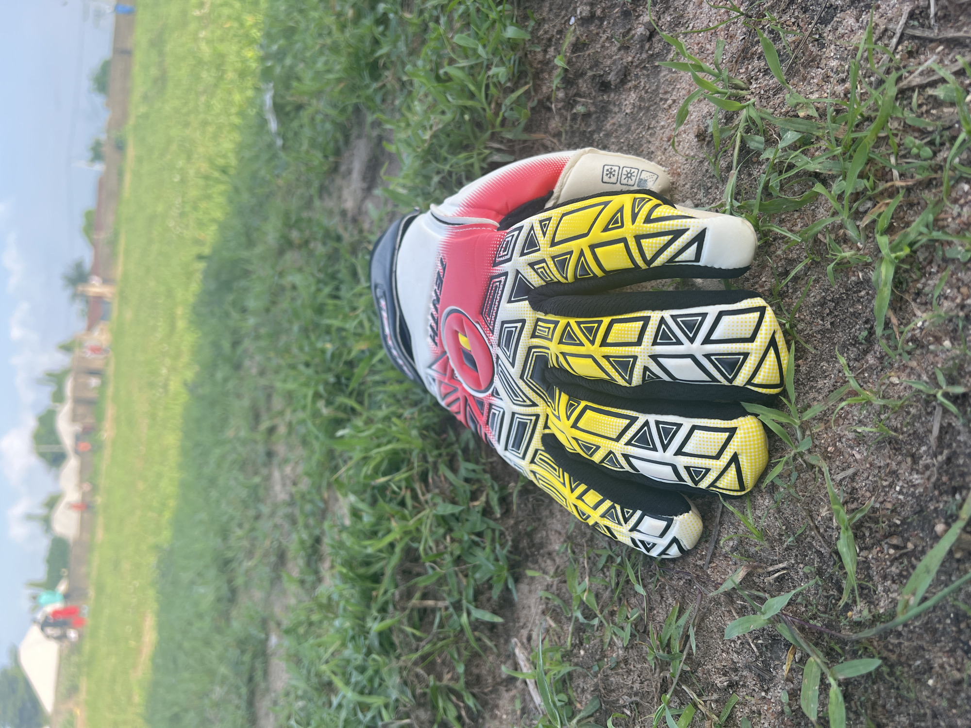 A single soccer goalkeeper glove lies on a patch of dry earth and short green grass, in an open field. The glove is predominantly yellow with black and white geometric patterns on the fingers and palm. Red and white accents are visible on the backhand, along with black wrist closure. In the background, a blurred landscape shows a low brick wall and several buildings with white, conical roofs, possibly tents or shelters. A few indistinct figures, appearing as small, blurred shapes, are scattered across the field in the distance. The sky is bright and overcast. The scene is likely located in Jalingo, Nigeria, given the context.