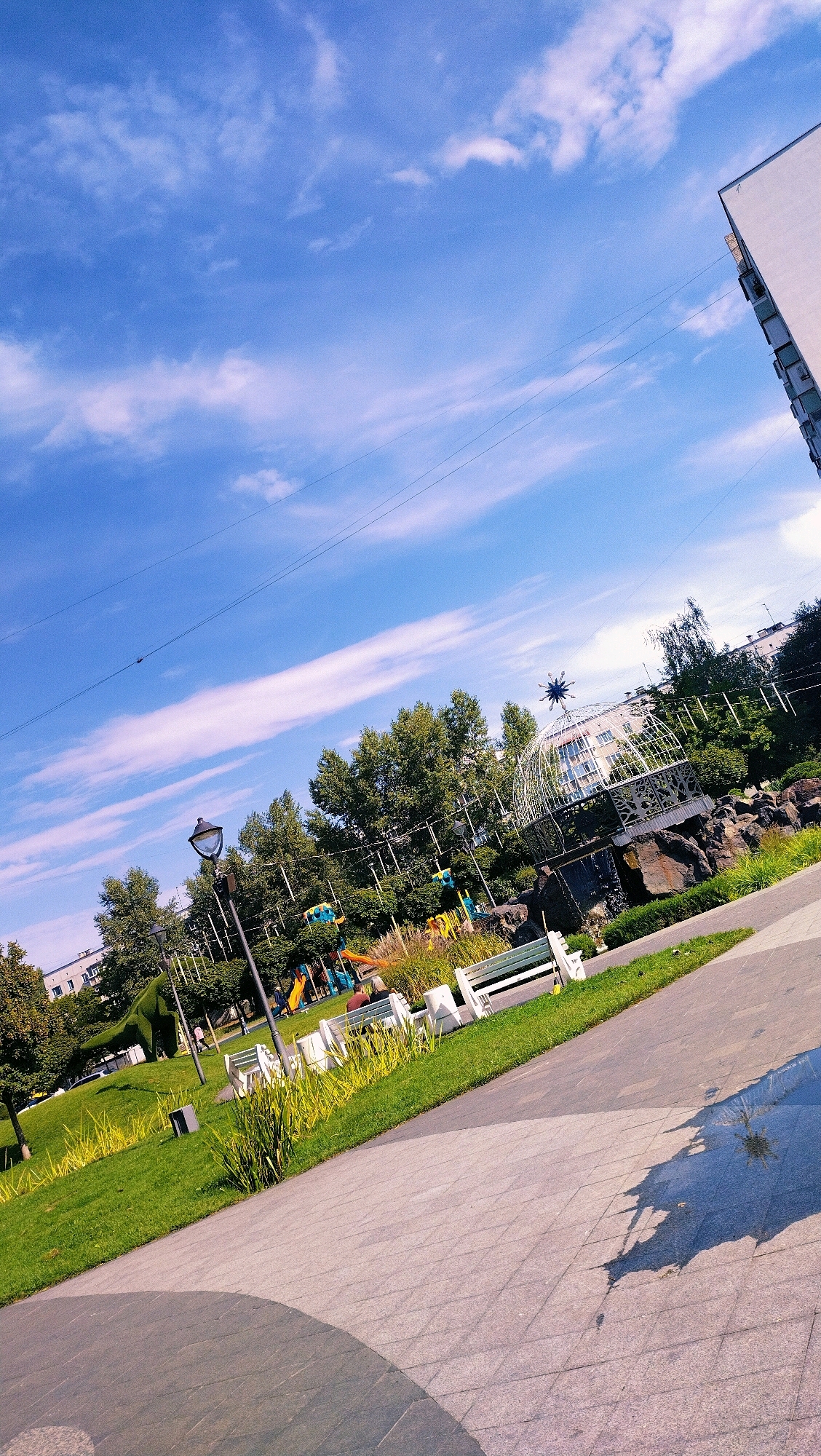 The image depicts a park area in Horishni Plavni, Ukraine, on a sunny day with a bright blue sky and scattered white clouds. The perspective is angled, giving a dynamic feel to the scene. In the foreground, a paved walkway made of gray stone tiles with a distinct pattern is visible. The tiles are arranged in a geometric pattern, and there is a circular shadow cast on the walkway, possibly from an overhead structure or tree.
To the left and extending into the midground, a well-maintained green lawn is present, with patches of taller, golden grass. Several white park benches are arranged along the edge of the walkway and on the lawn. One bench in the midground has two individuals seated on it, facing away from the camera. Another bench further back also has a single person seated.
Behind the benches and lawn, a children's playground is partially visible, with colorful structures like slides and climbing equipment. A tall lamppost stands in the midground, casting a shadow on the ground. Further into the background, a dense line of green trees forms a backdrop. Beyond the trees, several multi-story buildings with white facades are visible, indicating the urban setting.
A prominent architectural feature in the mid-to-background is a white, geodesic dome-like structure with a star-shaped element at its apex, situated on a rocky outcrop. A small waterfall is visible cascading down the rocks near this structure. Overhead, thin black wires, likely power or communication lines, crisscross the sky.
On the far right of the image, the corner of a modern white building with visible window frames and balconies juts into the frame from the top right, suggesting it's part of the urban landscape surrounding the park. The overall impression is a vibrant and well-kept public space within a city.