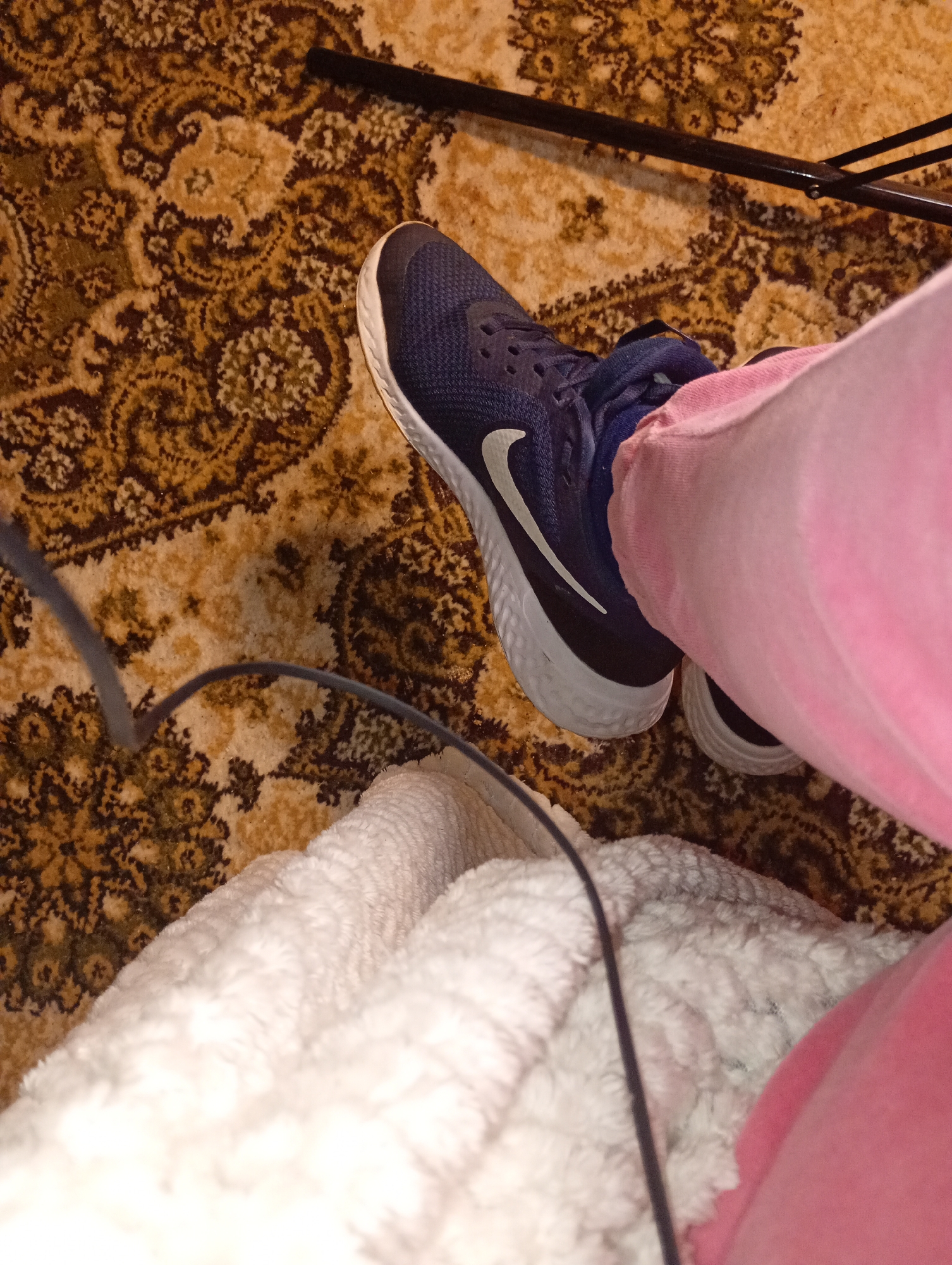 An overhead view depicts a segment of a brown and beige patterned carpet, featuring traditional motifs, within an indoor environment in Unknown, Unknown. A person's lower leg and foot are partially visible, clad in pink fabric, consistent with sweatpants, and a dark blue athletic shoe with a white Nike swoosh and a white textured sole. A black, slender, rigid object, possibly a furniture leg or pole, extends diagonally from the upper left towards the upper right. A dark gray cable traverses the lower left and central sections of the frame. A white, plush, textured blanket or throw partially covers the carpet in the lower portion of the image.