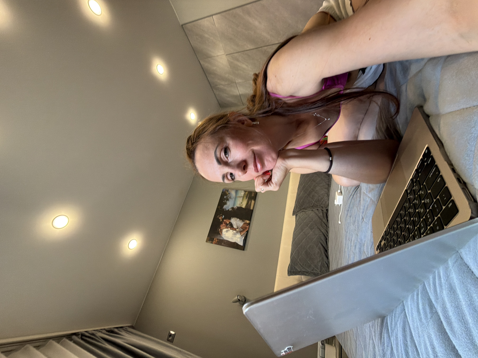 The image shows a woman lying on a bed with a laptop in front of her. She is looking at the camera and resting her chin on her hand. The room appears to be a bedroom with a gray wall, a framed picture, and ceiling lights. The woman is wearing a pink top and white shorts. The bed is covered with a blue textured blanket and gray pillows. A laptop, a charging adapter, and a curtain are also visible. The image does not contain any information that would suggest a location in Birobidzhan, Russia.