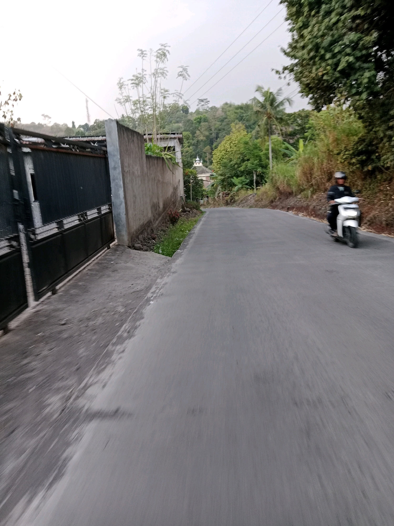 The image depicts a street scene in Sirnagalih, Indonesia. A person is riding a white scooter on a paved road that curves to the right. The scooter rider is wearing a helmet and a dark jacket. To the left of the road is a concrete wall and a black metal fence. Beyond the fence, there are some tall plants. The background features lush green hills covered in trees. The sky is overcast. The overall impression is a rural or suburban setting with a clear sense of motion due to the perspective and slight blur.