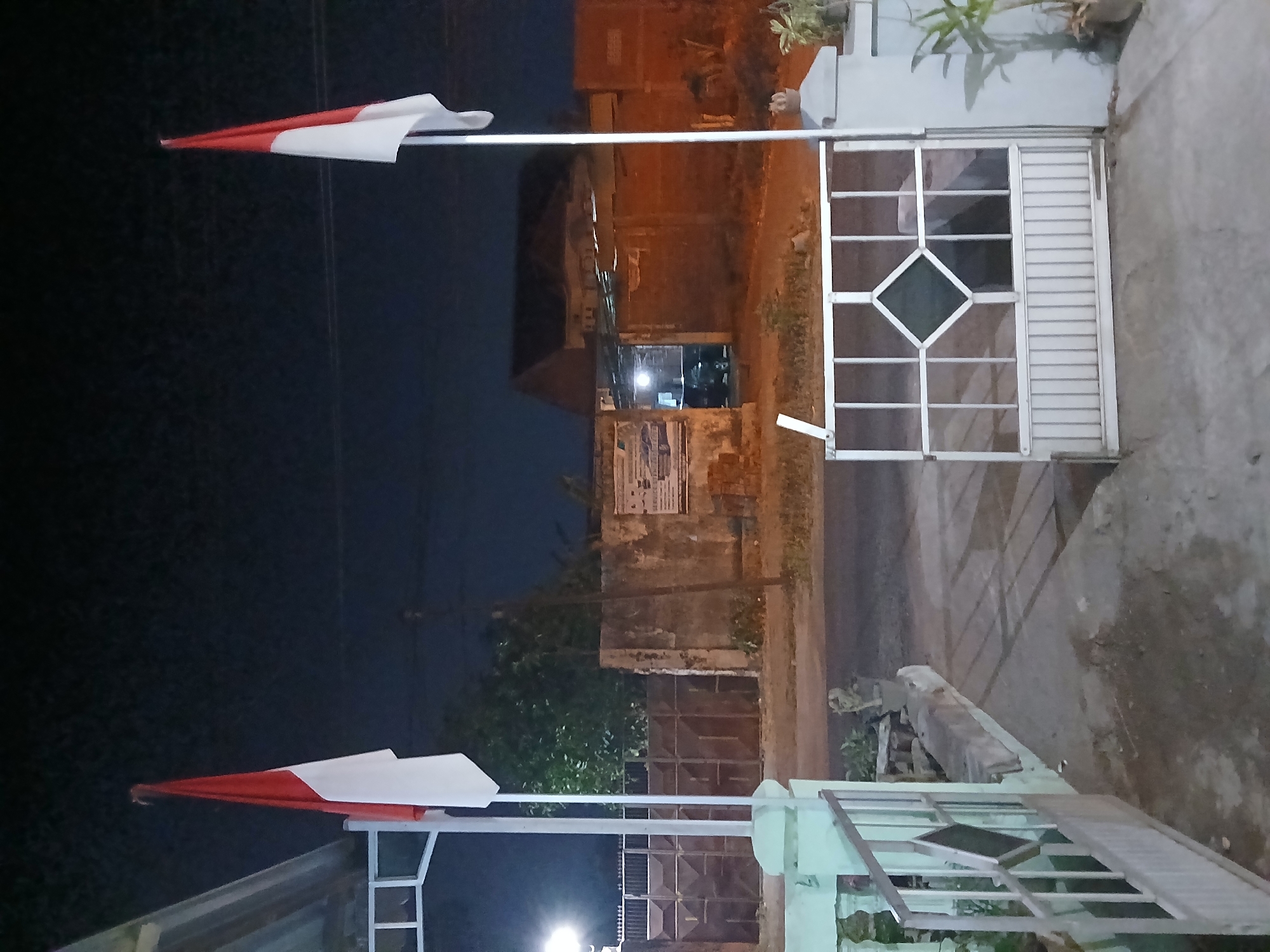 The image shows a nighttime view of two Indonesian flags, each displayed on a flagpole. The flag on the left is red and white, with the red portion at the top. The flag on the right is also red and white, positioned in a similar orientation. Both flags appear to be attached to poles. In the background, there is a building with a dark roof and illuminated windows. There are also fences and a gate. The overall environment suggests an outdoor setting at night. The location is identified as Kademangan Kulon, Indonesia.