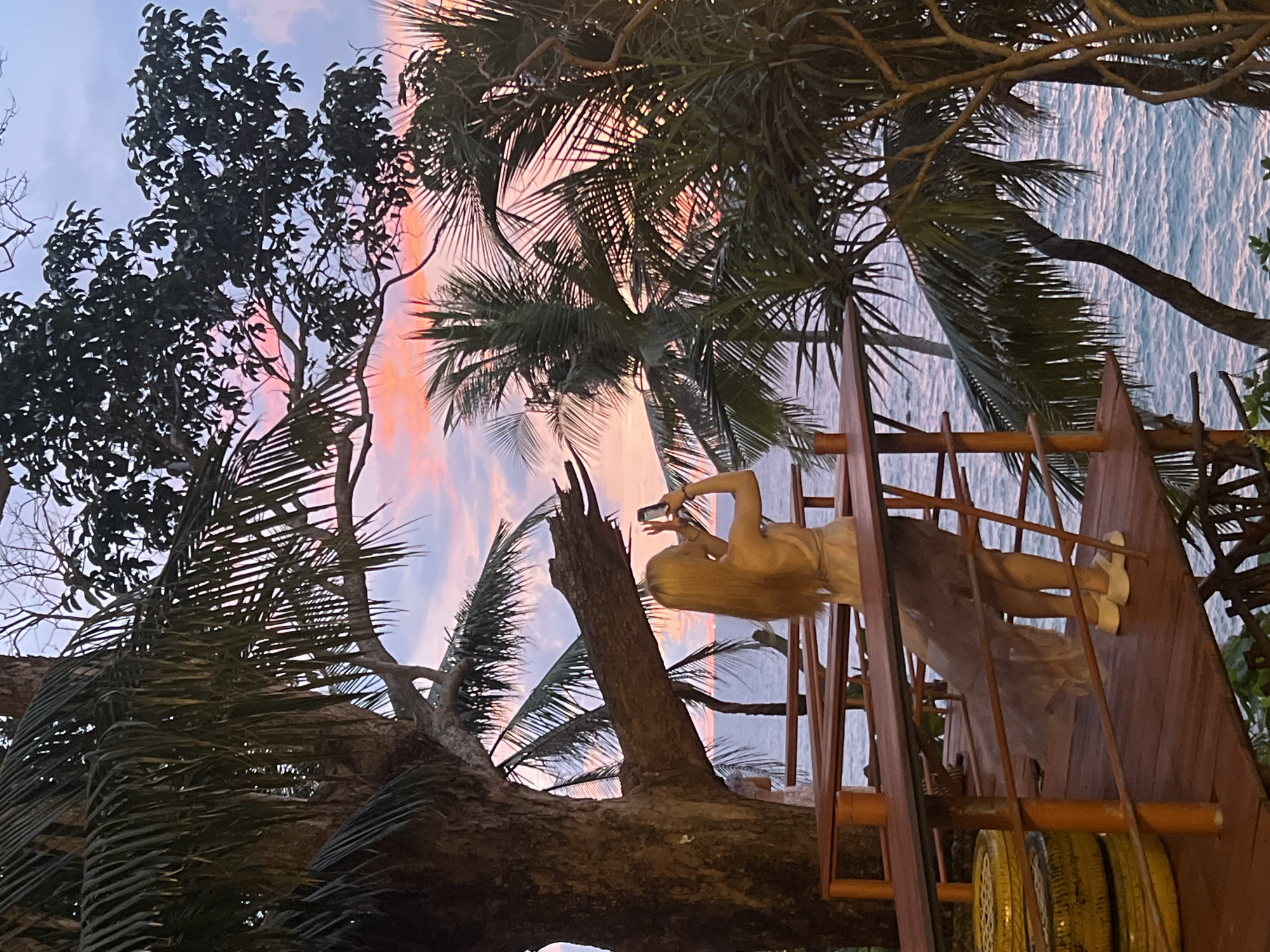 A woman with long blonde hair stands on a wooden platform overlooking the ocean at sunset. She is wearing a light-colored dress and white sandals and is holding a smartphone up to take a picture. Palm trees frame the scene, with their fronds silhouetted against a sky painted with hues of pink, orange, and purple. The ocean waves are visible below, reflecting the colors of the sunset. The platform appears to be part of an elevated structure, possibly a treehouse or a restaurant deck, with wooden railings and supports. In the foreground, stacked yellow tires are visible, suggesting a rustic or bohemian setting. The overall ambiance is serene and picturesque, capturing a moment of leisure and appreciation for the natural beauty of Karon, Thailand.