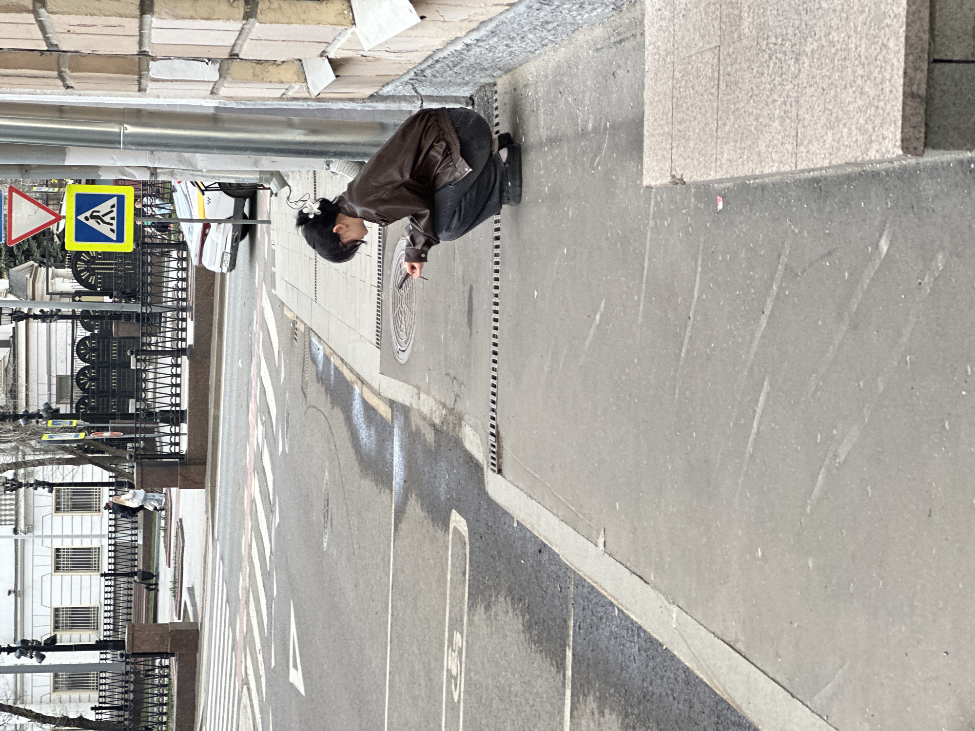 The image depicts a street scene in Moscow, Russia. A person is squatting near the curb of the street. They are wearing a brown jacket, dark pants, and dark shoes with white socks. They have dark hair. The person appears to be looking at something on the ground. They are holding a small cylindrical object.
A pedestrian crossing sign is visible on the left side of the street. A building with numerous windows and a black fence is visible in the background. The street is wet, suggesting it may have recently rained.