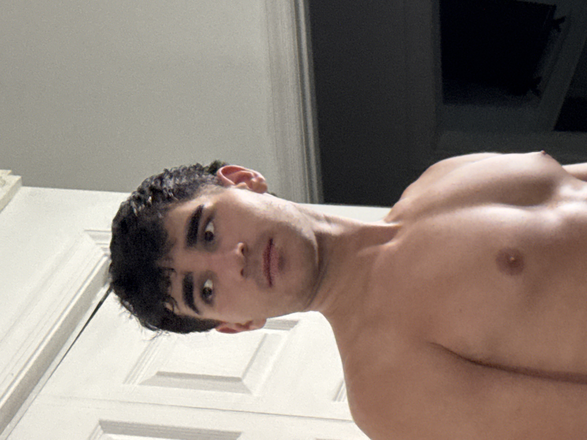 The image shows a young, shirtless man with dark, curly hair, looking to his right. He has dark eyebrows and eyes. He is standing in an indoor setting, with a white door to his left and a gray wall and a dark television screen in the background. The lighting appears to be artificial, creating shadows on his chest and shoulders. There is a white molding or trim visible at the top of the image. The overall scene suggests a casual or personal moment captured in what appears to be a residential environment within Bellmore, New York.
