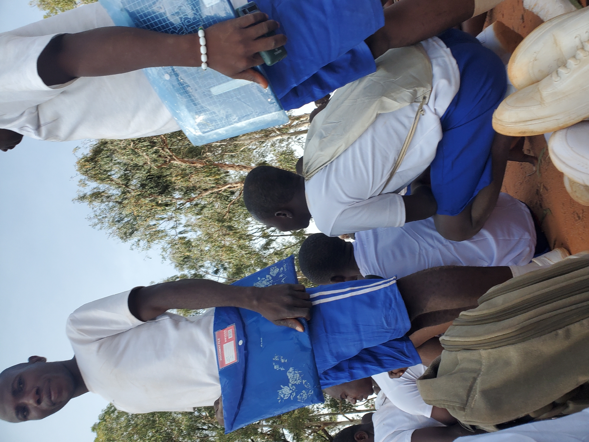The image depicts a group of young men, likely students, gathered outdoors in what appears to be a dusty, open area, possibly a schoolyard or training ground in Barakin Rafin Gora, Nigeria. The lighting suggests it is daytime, with a bright sky. The men are predominantly dressed in white t-shirts and blue shorts.
In the foreground, a young man is prominently featured, facing towards the camera with his head slightly tilted. He is wearing a white t-shirt and blue shorts with white stripes along the side. He is holding a blue document folder with floral patterns and text that appears to read "DOCUMENT FILE." His right hand, adorned with a white beaded bracelet, is resting on the folder.
To his left, another young man in a white t-shirt and blue shorts is partially visible, looking off to the side. Further to the left, a duffel bag, a light brown or olive color, is placed on the ground.
In the mid-ground, another individual, also in a white t-shirt and blue shorts, is seen from behind, crouched down. A light-colored cloth or bag is draped over his shoulders. Adjacent to him, to his right, a portion of another person wearing a white t-shirt and blue shorts is visible.
The background features trees with sparse foliage, indicating a warm climate. The ground is covered in reddish-brown dirt. The overall impression is one of an organized gathering, possibly a school event, orientation, or a practical training session. The presence of document folders and the uniform attire suggest an official capacity.