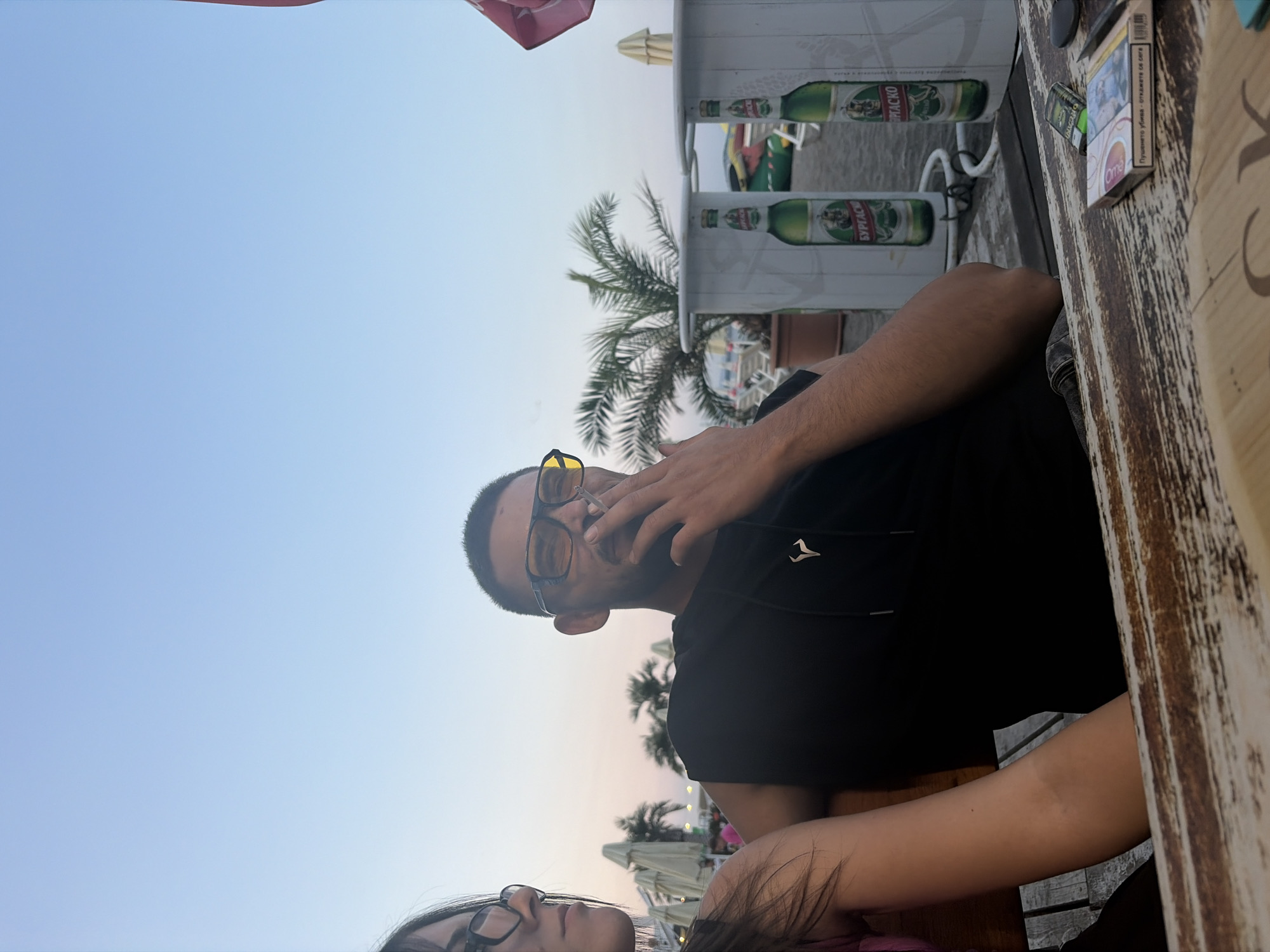 A vertically oriented image shows two people at an outdoor establishment, possibly a beach cafe, in Burgas, Bulgaria. The sky suggests it is late afternoon or early evening, with a soft gradient of blue and pink hues. In the foreground, a man with glasses and a black hoodie is seen from the chest up, with his right hand near his mouth, appearing to smoke. His gaze is directed towards the camera. To the left and below him, a woman is partially visible, also wearing glasses and looking towards the man.
Behind them, a wooden table is visible, with a pack of cigarettes and a lighter on its surface. Further in the background, a white structure with images of green beer bottles and text is present, possibly part of a bar or display. Palm trees and other beach umbrellas are scattered in the background, indicating a warm, coastal environment. The overall mood seems relaxed and casual. The visible text on the beer bottles appears to be in Cyrillic.
