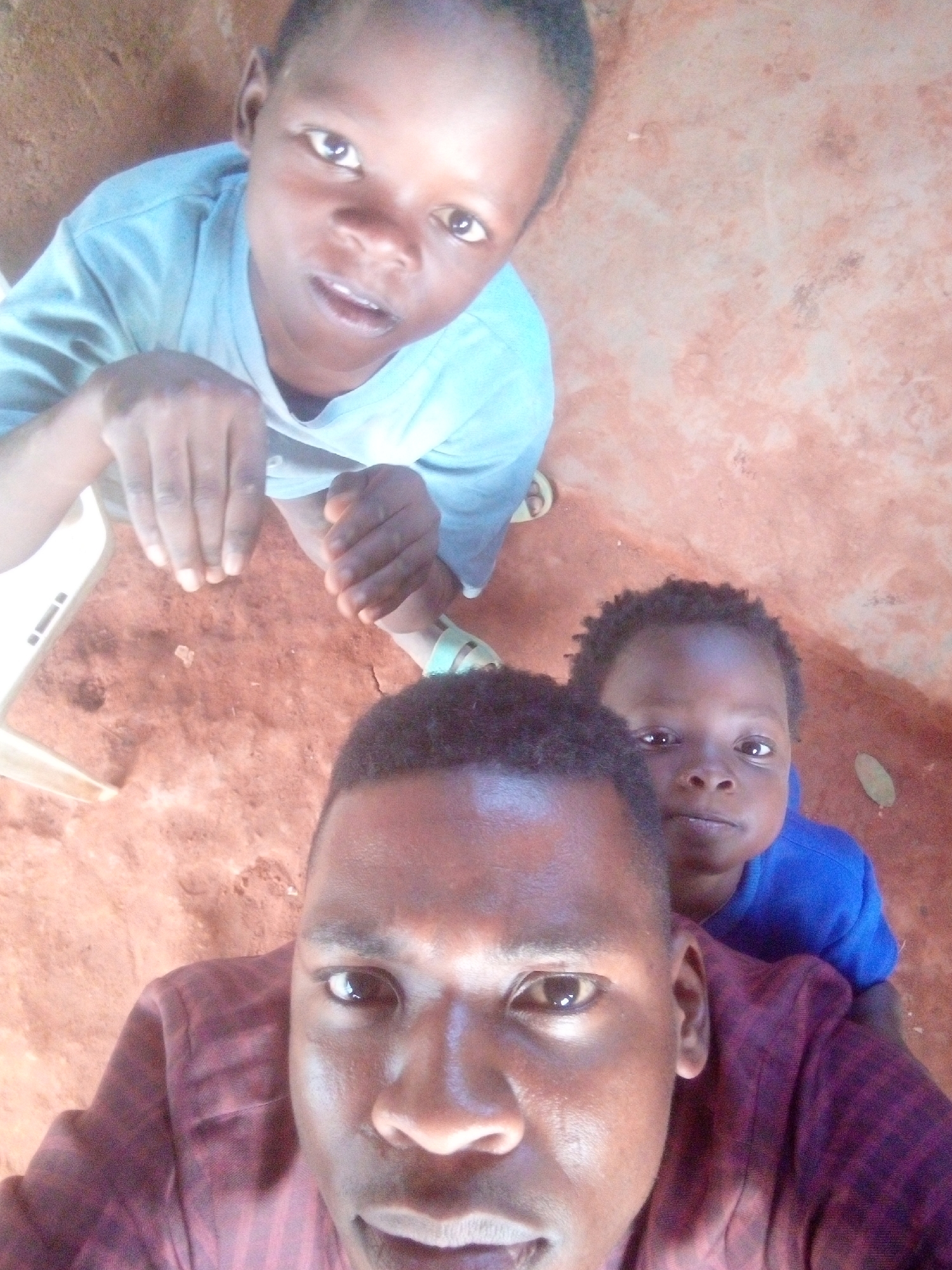The image shows a selfie taken from a low angle, featuring a dark-skinned man in the foreground and two children in the background. The man is wearing a dark red shirt and has short, dark hair. The child in the top left corner is wearing a light blue shirt and looking directly at the camera with their hands visible. The child to the right of the man is wearing a blue shirt and looking forward. The location appears to be an indoor or covered area with a reddish-brown floor. The specific city or region is not identifiable.
