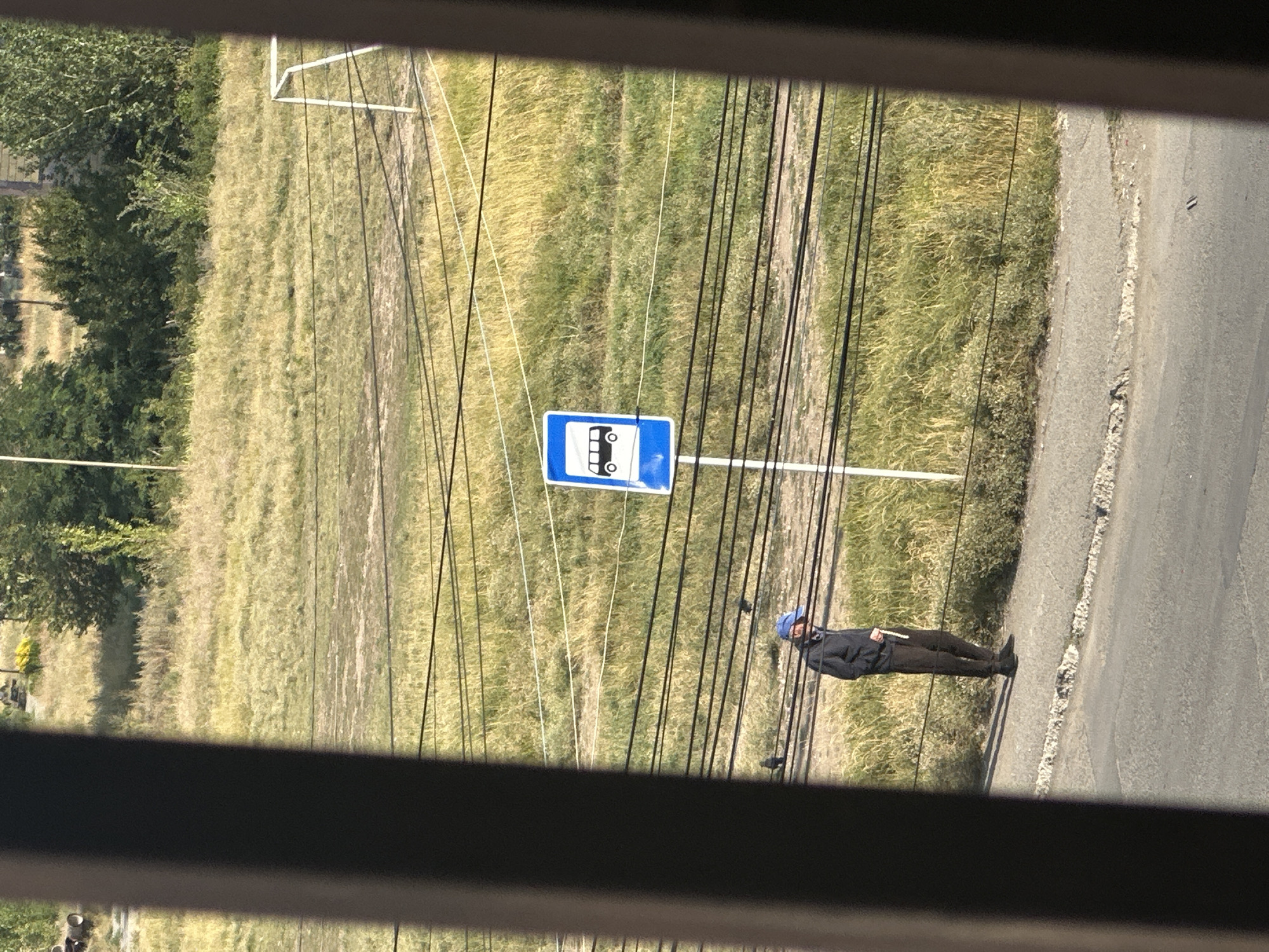 A full-shot, medium-distance photograph depicts a man standing on a sidewalk next to a bus stop sign. He is wearing a dark jacket, dark pants, and a blue baseball cap. The bus stop sign is blue and white, with a pictogram of a bus. The man is holding a thin, white object in his right hand. Behind the man and the bus stop, there is a grassy field with some trees in the background. Several black wires are strung horizontally across the scene. In the far background, a white soccer goal frame is visible. The scene appears to be an outdoor public area in Gyumri, Armenia, likely on the outskirts of the city, given the presence of the field and wires. The lighting suggests it is daytime, possibly late afternoon due to the shadows.