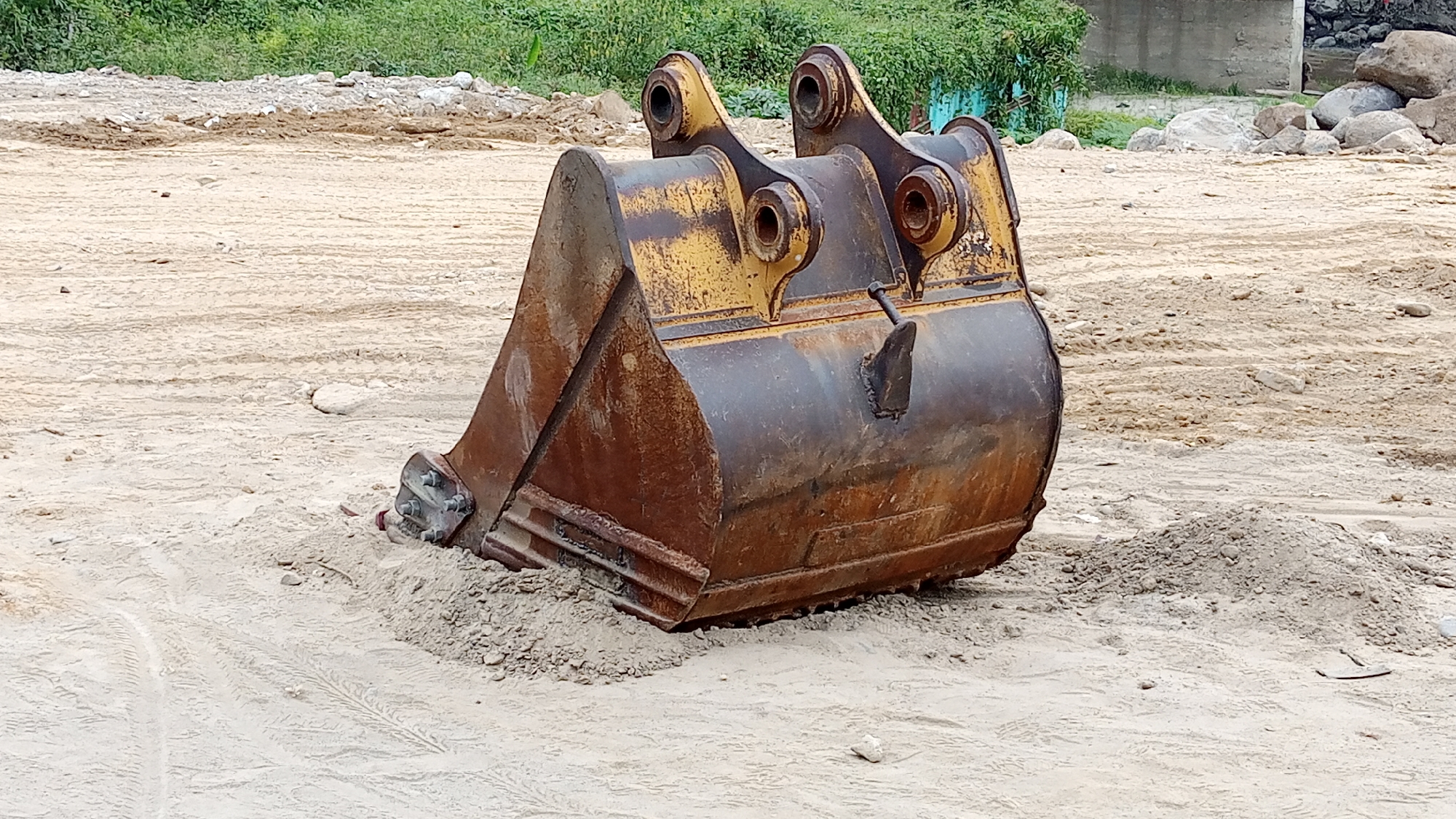 An excavator bucket is partially submerged in loose sand. The bucket is made of rusted metal with yellow paint markings. It has a wide, angled opening with several teeth or blades along the bottom edge. Two large metal pins connect the bucket to a pivot mechanism at the top, which is also yellow and appears worn. The bucket is resting on a patch of granular material, possibly gravel or coarse sand. The surrounding environment is a construction or undeveloped area, characterized by large expanses of light-colored sand with tire tracks visible in places. In the background, there are piles of dirt and debris, some scattered rocks, and a dense line of green vegetation. A concrete structure is partially visible in the far background. There are no people or animals present in the image. The scene appears to be outdoors during daylight. The location is identified as Beunyot, Indonesia.