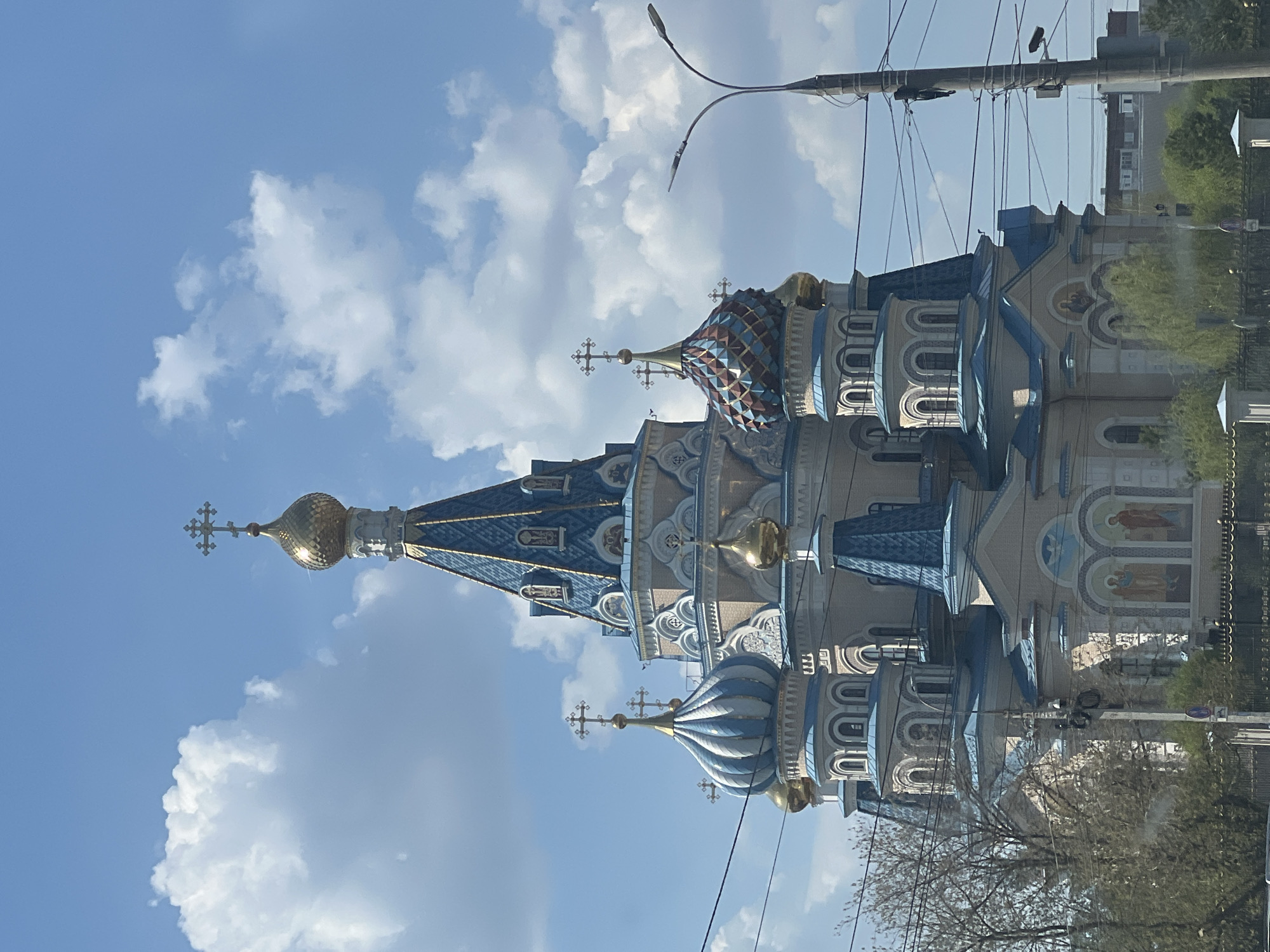 The image depicts the Alexander Nevsky Cathedral in Izhevsk, Russia. The cathedral features multiple domes with varying colors, including gold, blue, and a patterned red, blue, and tan. The building is light-colored with blue and white architectural details. The sky is blue with puffy white clouds. Power lines are visible in the foreground, running diagonally across the image. Street lights and a few trees are also visible.