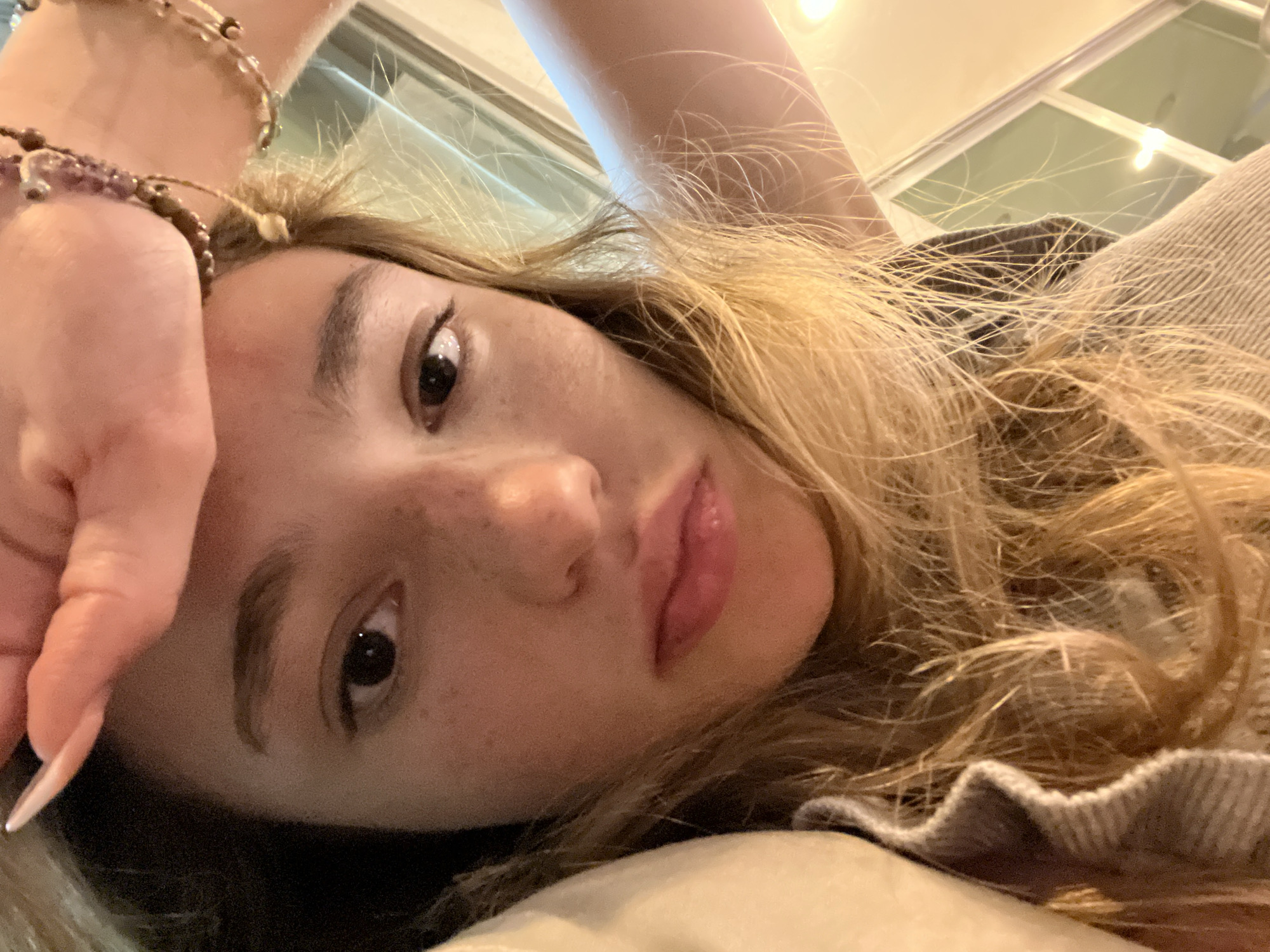 A close-up, angled selfie shows a young woman with fair skin, freckles, and light brown hair. Her right hand, adorned with beaded bracelets, is held near her forehead. She has dark eyes, and her lips are slightly parted. She is wearing a gray sweater with a ruffled collar. The background suggests an indoor setting with a window visible. There is no information to connect the picture to the city of San Bernardino Tlaxcalancingo, Mexico.