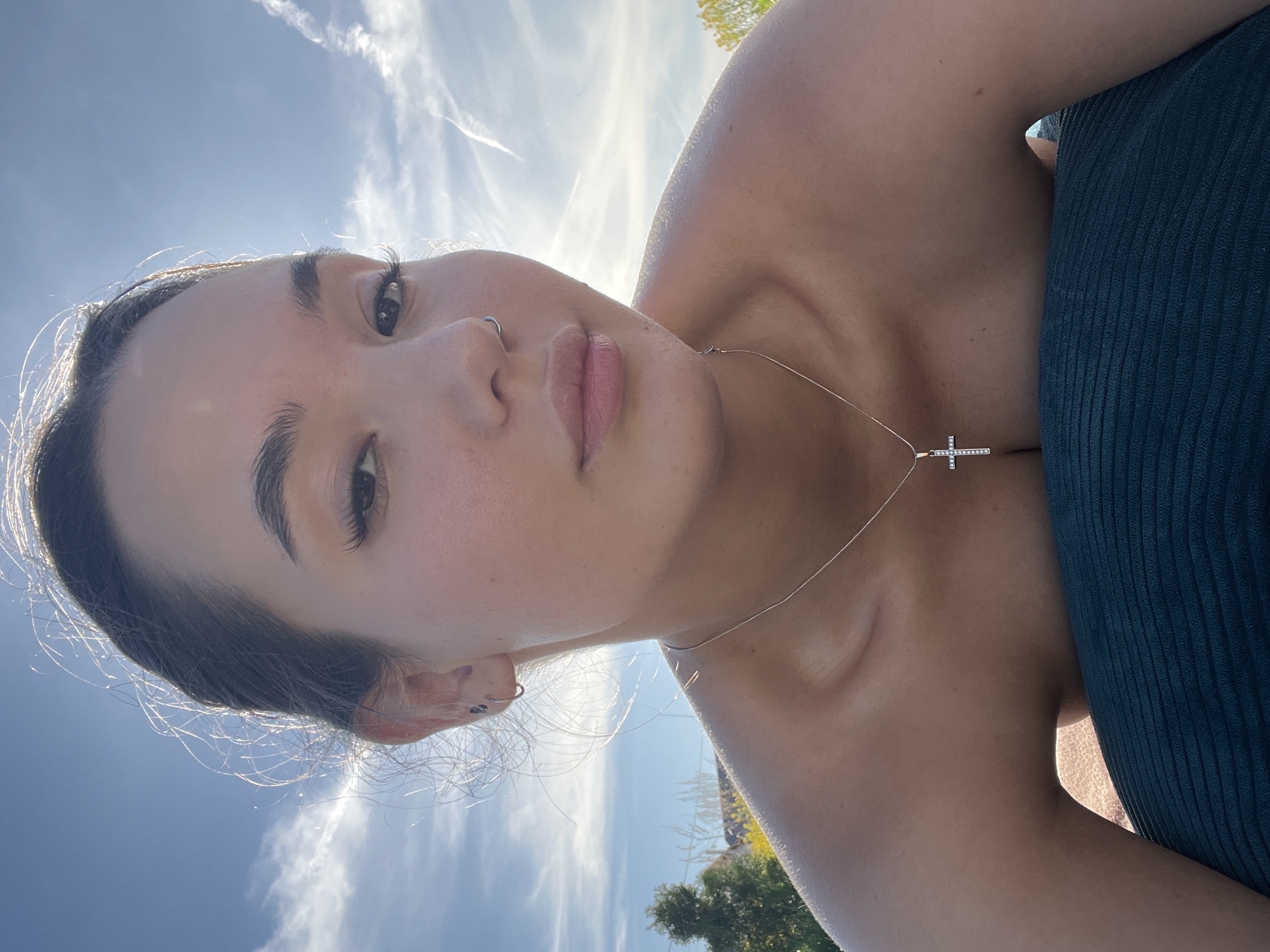 A close-up, rotated view of a light-skinned woman. She has dark hair pulled back, dark eyebrows, eyelashes, a nose piercing on the left side, and is wearing a necklace with a cross pendant. The background shows a blue sky with light clouds. She appears to be wearing a dark green top. The location is assumed to be near Лобаново, Russia, based on the context.