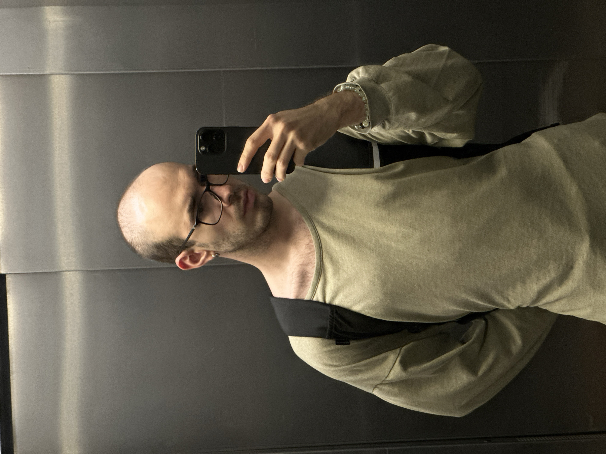 A man is taking a selfie in an elevator. The man has a shaved head, wears glasses, a light green shirt, and a pearl bracelet. He is holding a black smartphone. A black strap, likely from a bag, is visible over his shoulder. The elevator has brushed metal walls. The location of the scene is not in the city of Khimki, Russia.