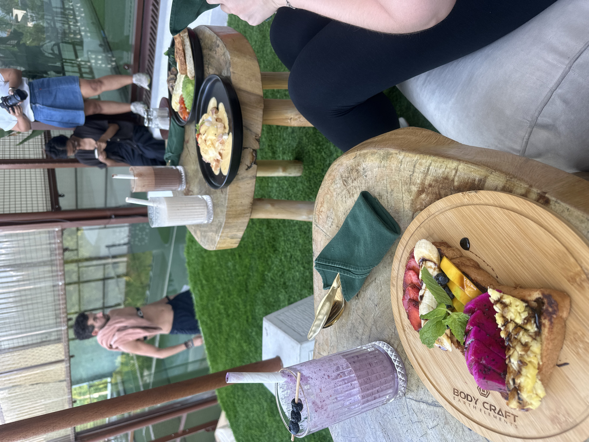 An outdoor setting with artificial grass and wooden furniture. A man with dark hair is shirtless with a pink towel draped over his shoulders, wearing dark shorts. He is standing in the background. In the foreground, a wooden platter holds a slice of toast topped with scrambled eggs, dragon fruit, mango, banana, strawberries, and mint. A glass of purple berry smoothie with a paper straw and blueberries is next to the platter. A dark green napkin is folded with gold cutlery on a wooden table. Two other tables are visible, one with plates of food and drinks, and another with salt and pepper shakers. In the background, two women are seated and looking at their phones, and another woman stands holding a camera. The scene appears to be a cafe or restaurant.