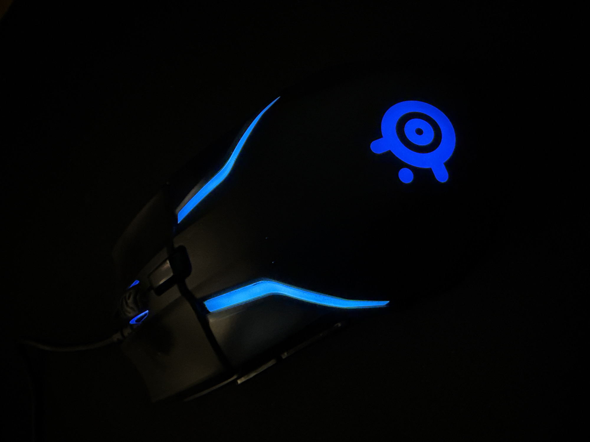 A black gaming mouse with blue LED lighting is positioned on a dark surface. The mouse has a sleek, ergonomic design with glowing blue accents along its sides and at the top near the scroll wheel. The SteelSeries logo, also illuminated in blue, is visible on the palm rest area of the mouse. The background is entirely black, indicating a dimly lit or dark environment. The image is taken from a top-down perspective, focusing on the mouse. There is no discernible location in the city of Mariannelund, Sweden mentioned or visible in the image.