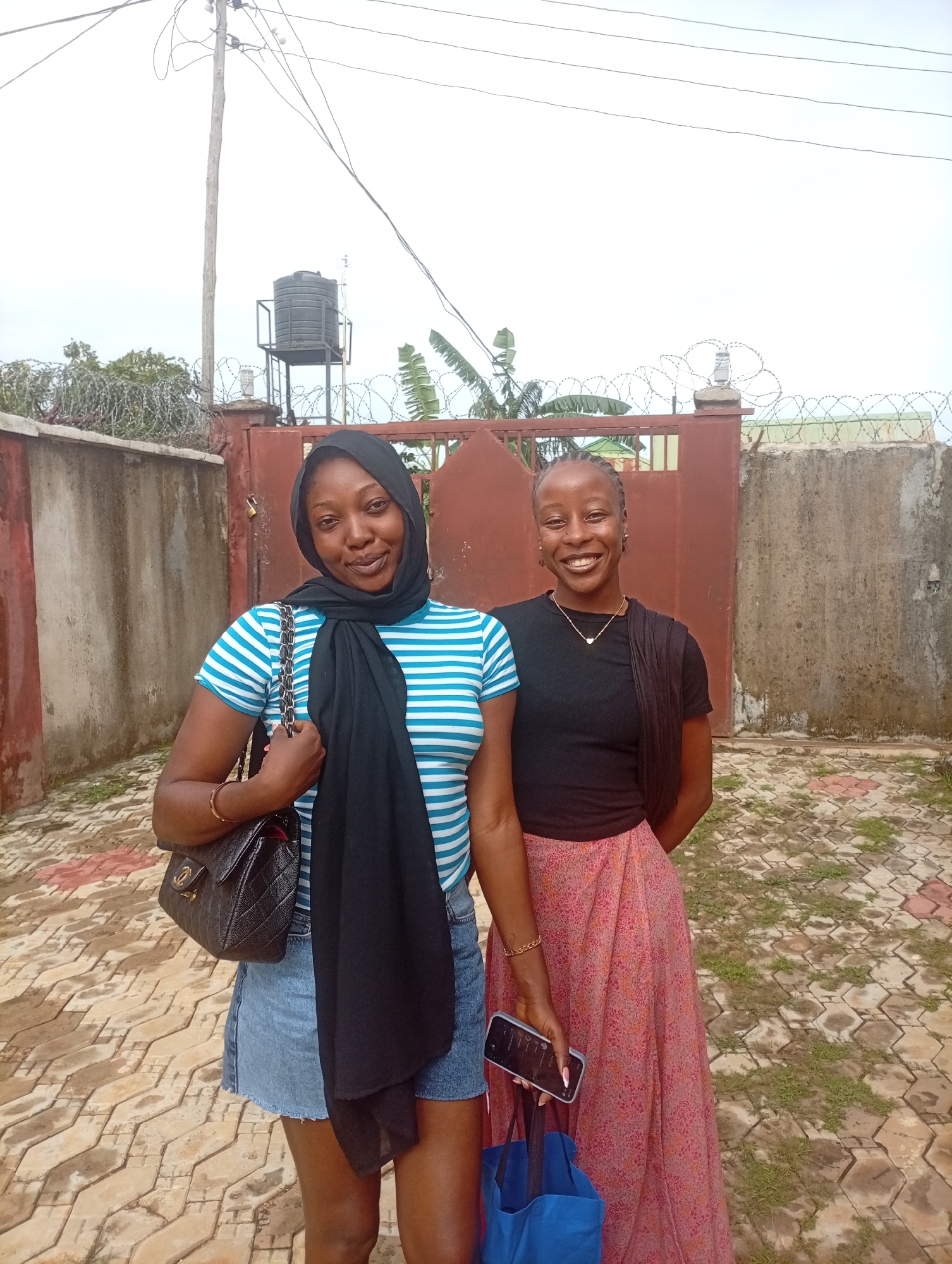 Two young Black women are standing in front of a red metal gate and a concrete wall topped with barbed wire. The woman on the left wears a blue and white striped t-shirt, denim shorts, a black hijab, and a black quilted handbag. The woman on the right wears a black t-shirt and a long, patterned pink skirt, and is holding a black smartphone and a blue tote bag. The ground is paved with interlocking hexagonal tiles. Behind the women, a water tank on a metal stand is visible, along with electrical wires. The sky is overcast.