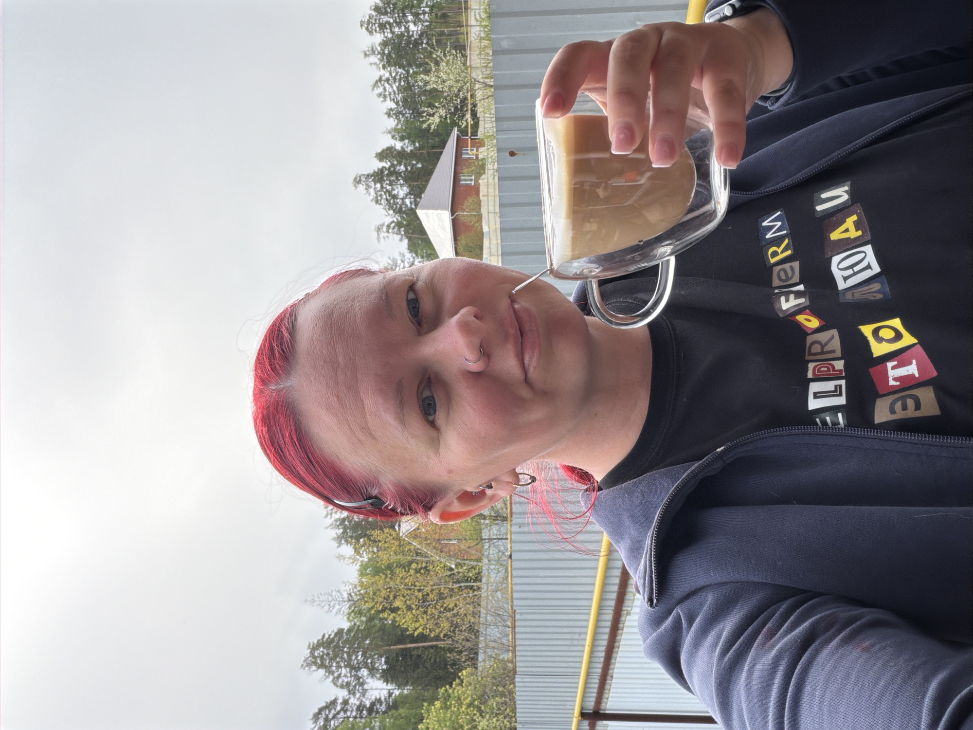 The image shows a selfie of a person with red hair and a nose ring, holding a clear glass mug filled with a coffee-like beverage. The person is wearing a black t-shirt with colorful lettering and a dark blue zip-up hoodie. The background shows a gray sky, trees, and a building. The location is Chaikovsky, Russia.