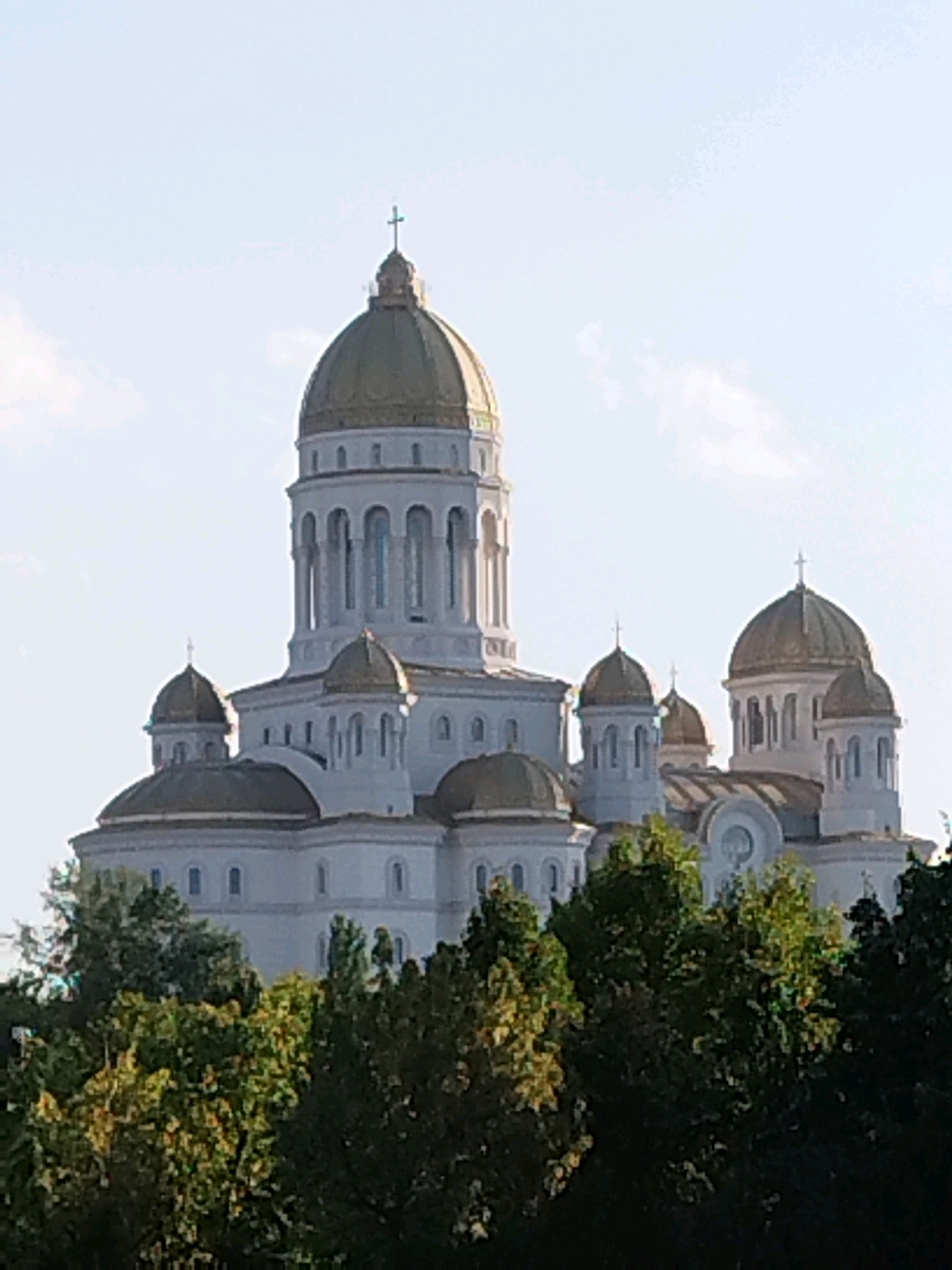 The image shows the Romanian Orthodox Cathedral in Bucharest, Romania. The cathedral is a large, white building with golden domes, set against a clear blue sky. It is surrounded by lush green trees. The cathedral is a prominent landmark in the city and is a popular tourist attraction.