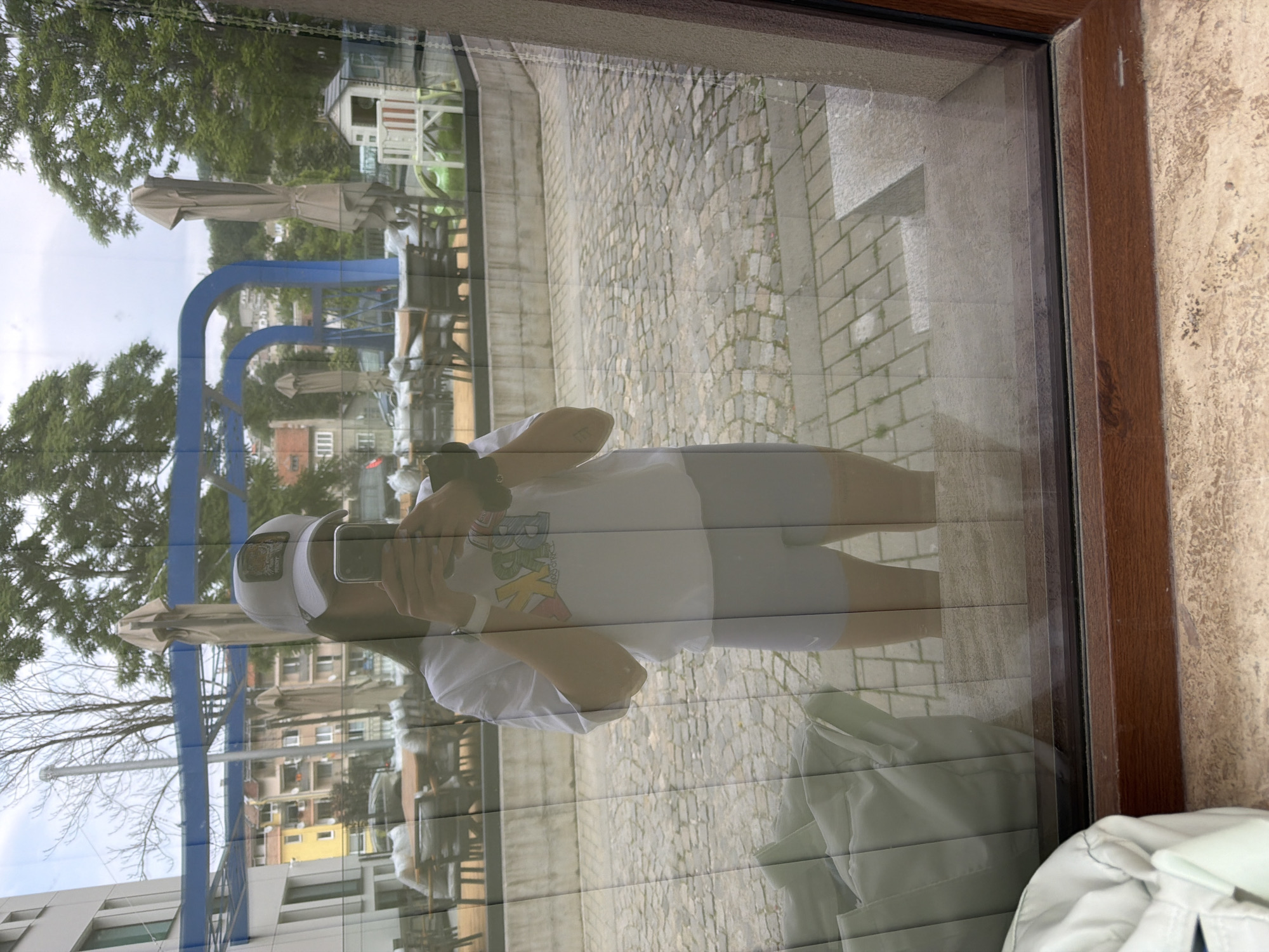 The image displays a reflection of a person taking a selfie in a glass window, revealing both the indoor foreground and the outdoor scene beyond.
The person, positioned centrally in the reflection, is wearing a white short-sleeved t-shirt with a multi-colored graphic design on the chest, light grey athletic shorts, and a light-colored baseball cap featuring a dark emblem, possibly an animal, on the front. A dark-haired individual is visible under the cap, holding a dark-colored smartphone with both hands to take the picture. A white smartwatch is visible on their left wrist.
Reflected and visible through the glass is an outdoor seating area with multiple tables and chairs, suggesting a cafe or restaurant patio. Some tables are covered with light-colored materials. Several large, closed beige umbrellas are interspersed among the tables. Beyond the patio, a cobblestone-paved ground extends. In the mid-ground, distinctive blue arched structures are visible, possibly part of a decorative or functional architectural element. To the right, a small white playhouse with a red roof, a green plastic slide or rocking toy, and a white child's chair are present.
The background features various buildings, including a modern, light-colored multi-story building on the far left, and older residential structures with varied facades (yellow, brown, white) further back. Lush green trees are abundant, particularly on the right and in the distant background, with some bare trees visible in the upper left quadrant. The sky is overcast.
In the immediate foreground, part of a light green bag is visible on an interior ledge to the left. The bottom of the image shows a dark brown wooden window frame and a light-colored, textured stone or concrete sill or ledge.
The scene is located in the city of Gabrovo, Bulgaria.