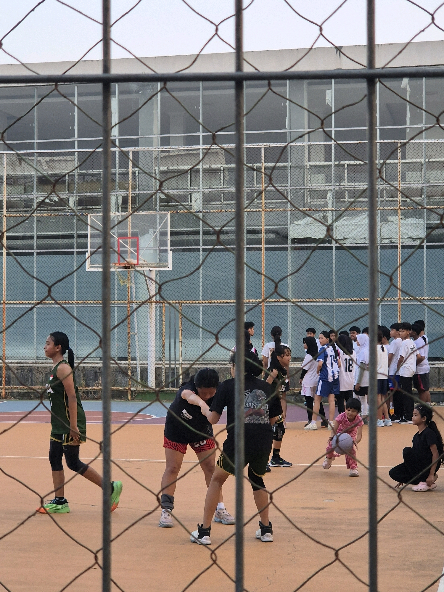 The image depicts a group of young people playing basketball on an outdoor court, viewed through a chain-link fence. The court has an orange surface with blue and red boundaries. A basketball hoop with a transparent backboard and a red rim is visible in the background. The individuals are dressed in athletic wear. One person in the foreground on the left is wearing a dark green tank top and black shorts with green and orange athletic shoes. Another person in the center foreground is wearing a black t-shirt with red and black striped shorts and black and white athletic shoes. A young child in pink clothing is in the middle ground, holding a basketball. Other people are gathered around the court. The background features a building with a large glass facade and a chain-link fence. The sky is overcast. The scene is likely located at a public sports facility in Samarinda, Indonesia, given the context provided.