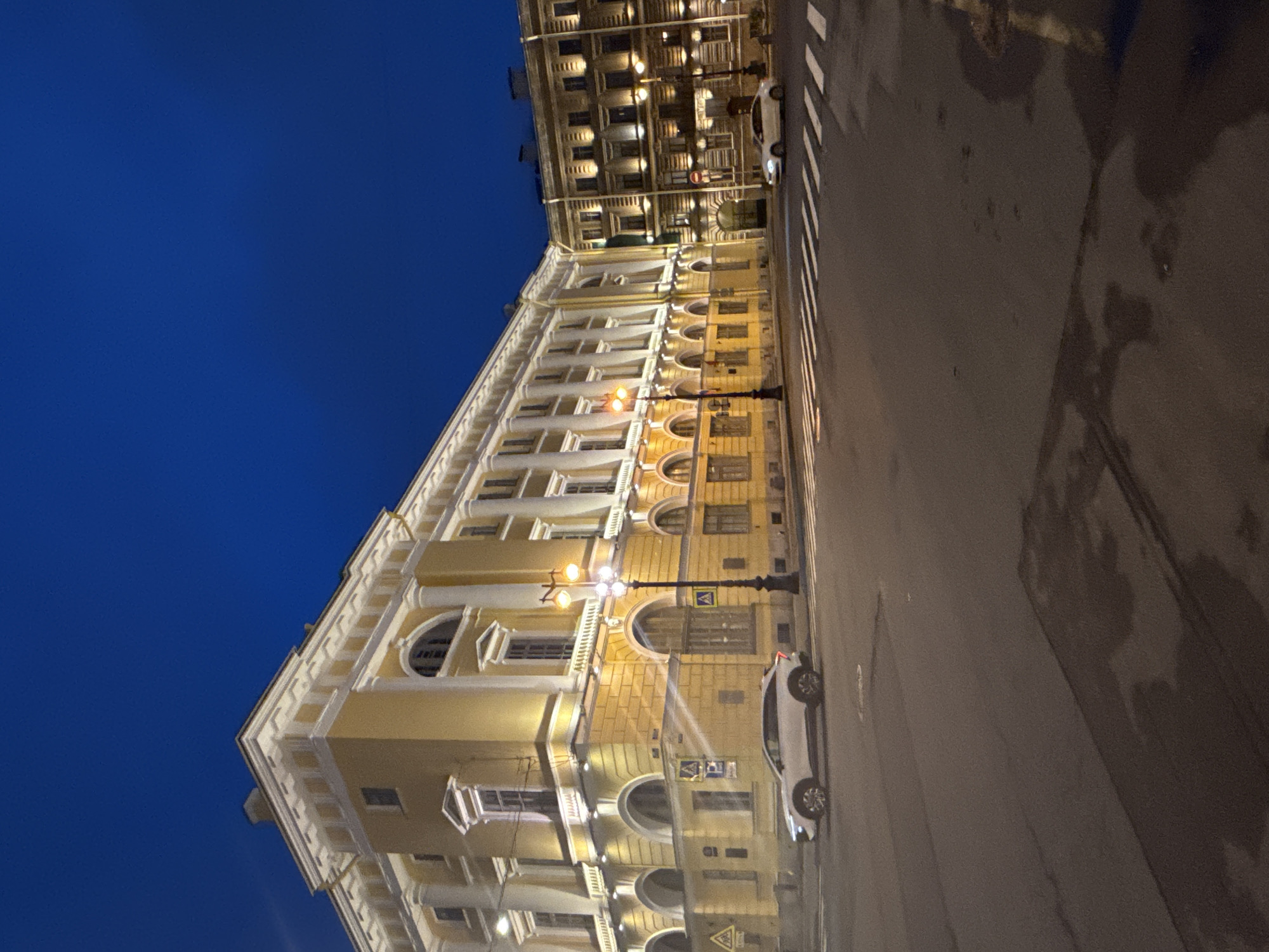 The image depicts a street view at night in Saint Petersburg, Russia, showcasing a predominantly yellow, multi-story building with ornate architectural details. The building has arched windows and decorative columns, illuminated by streetlights. A white car is parked along the side of the building, near a crosswalk. The sky is a deep blue.