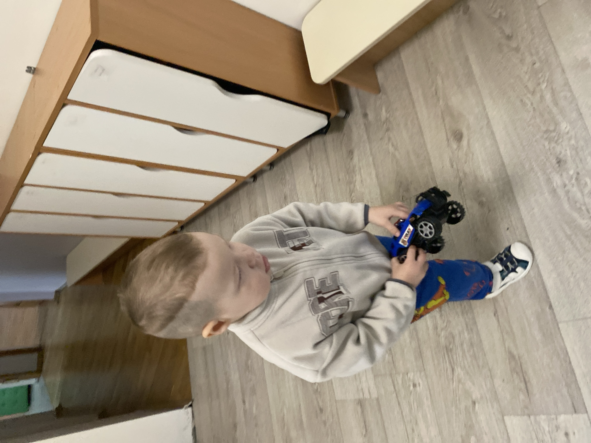 A young child stands on a light-colored, wood-patterned floor, holding a toy truck. The child wears a gray zippered sweatshirt with lettering on the front, blue patterned pants, and white sneakers with blue accents. To the left of the child is a low dresser with light brown wood and white drawers. Part of a table is visible above and to the right of the dresser. The scene's location is unknown, and there is no visible evidence to suggest it is in Sochi, Russia.