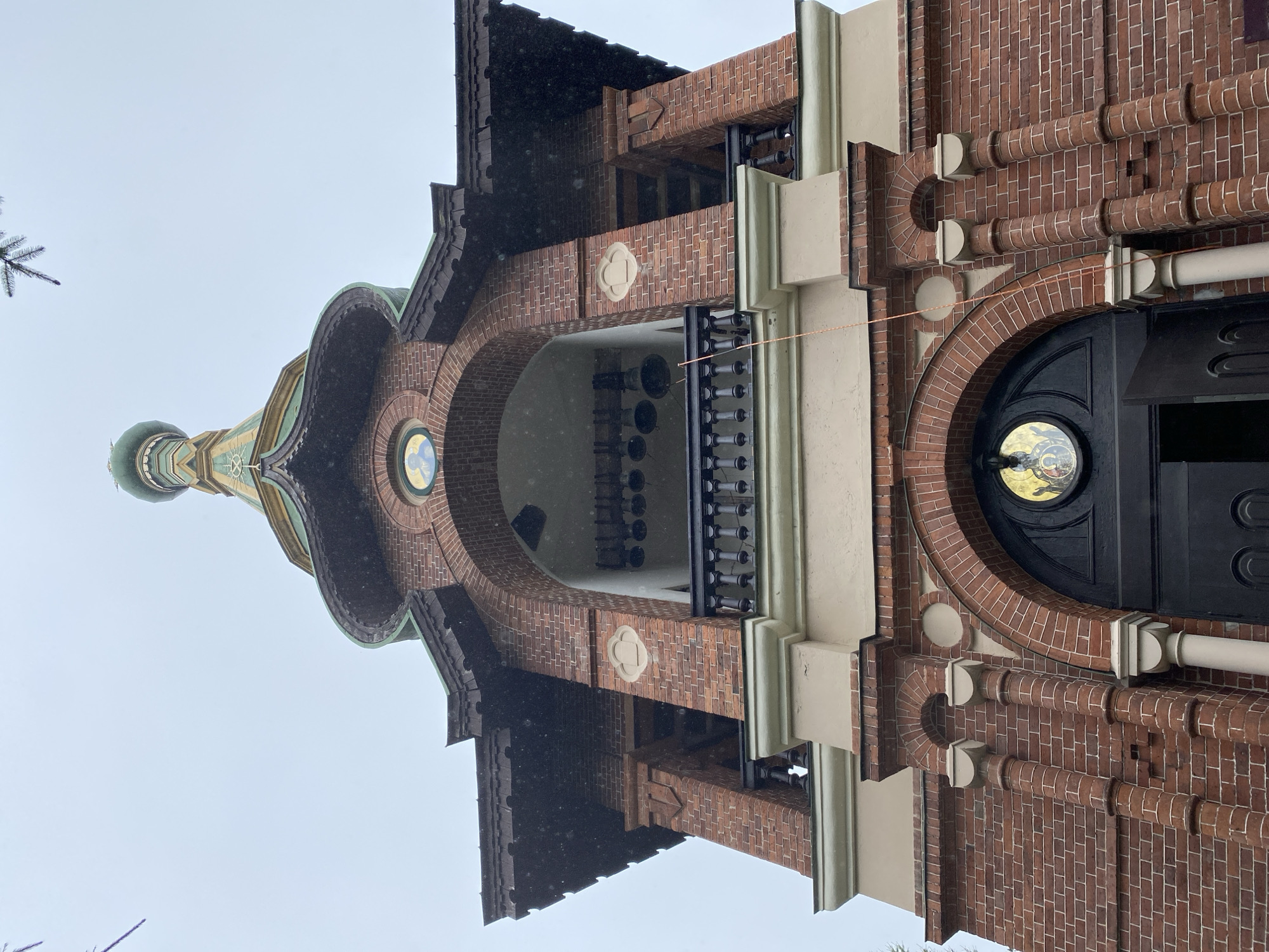 The image shows a low-angle view of the bell tower of the Church of the Transfiguration of the Lord in Priozersk, Russia. The tower is constructed of red brick with decorative architectural elements. It features a green, multi-tiered roof topped with a golden dome and cross. Arched openings reveal bells hanging inside, and a balcony with a black railing extends across the upper section. Below, a large dark wooden door is partially ajar. The sky is overcast, and a light snowfall is visible, with some evergreen branches extending into the frame from the top left. The scene is devoid of people.