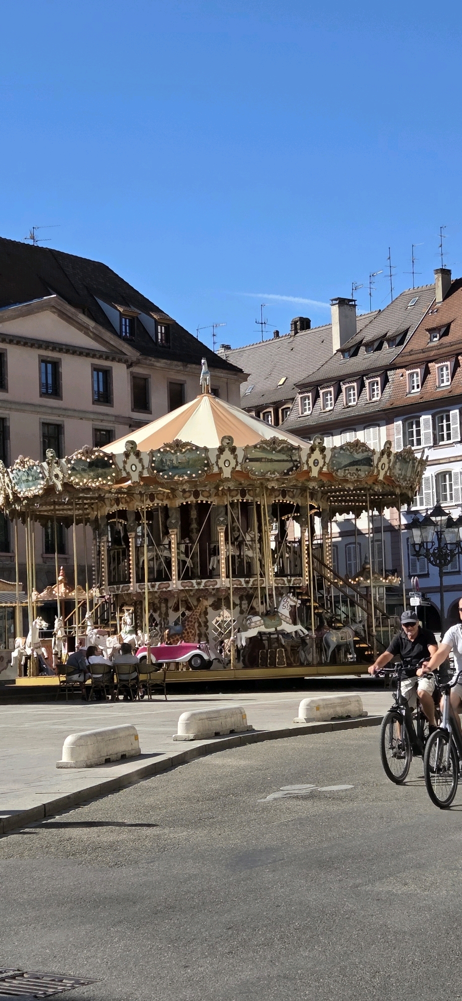 In Strasbourg, France, a classic carousel stands as the central attraction in a public square on a bright, clear day. The carousel is ornate, featuring various painted panels and multiple tiers of figures, including horses, a giraffe, and a vintage-style car. Several people are seated at tables set up in front of the carousel, suggesting a cafe or seating area. Two men are cycling in the foreground on the right side of the image, with one in focus and the other slightly blurred. The background consists of traditional European architecture with pitched roofs, dormer windows, and decorative facades. The overall scene conveys a lively and pleasant atmosphere in a European city.