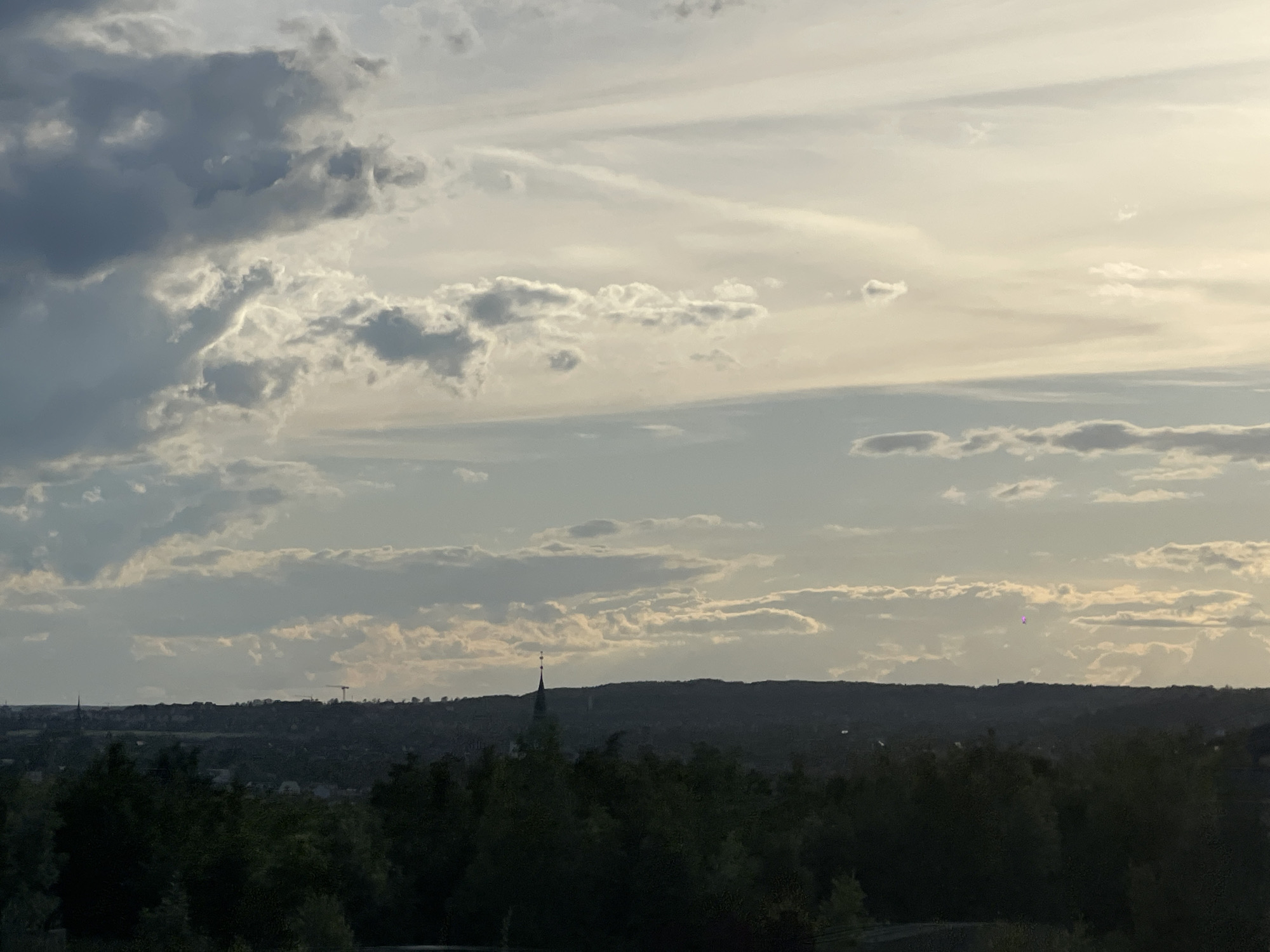 The image depicts a panoramic view of a city skyline at dusk, with dark, silhouetted trees in the foreground. The sky is filled with dramatic clouds, transitioning from dark, stormy grays on the left to lighter, wispy whites and yellows on the right, suggesting the last rays of sunlight. A thin, spired structure, possibly a church steeple, rises from the distant, hilly horizon. Farther to the right, a construction crane is visible, and a small, glowing purple object floats in the sky. The overall impression is one of a serene, yet slightly ominous, twilight scene over an urban landscape.