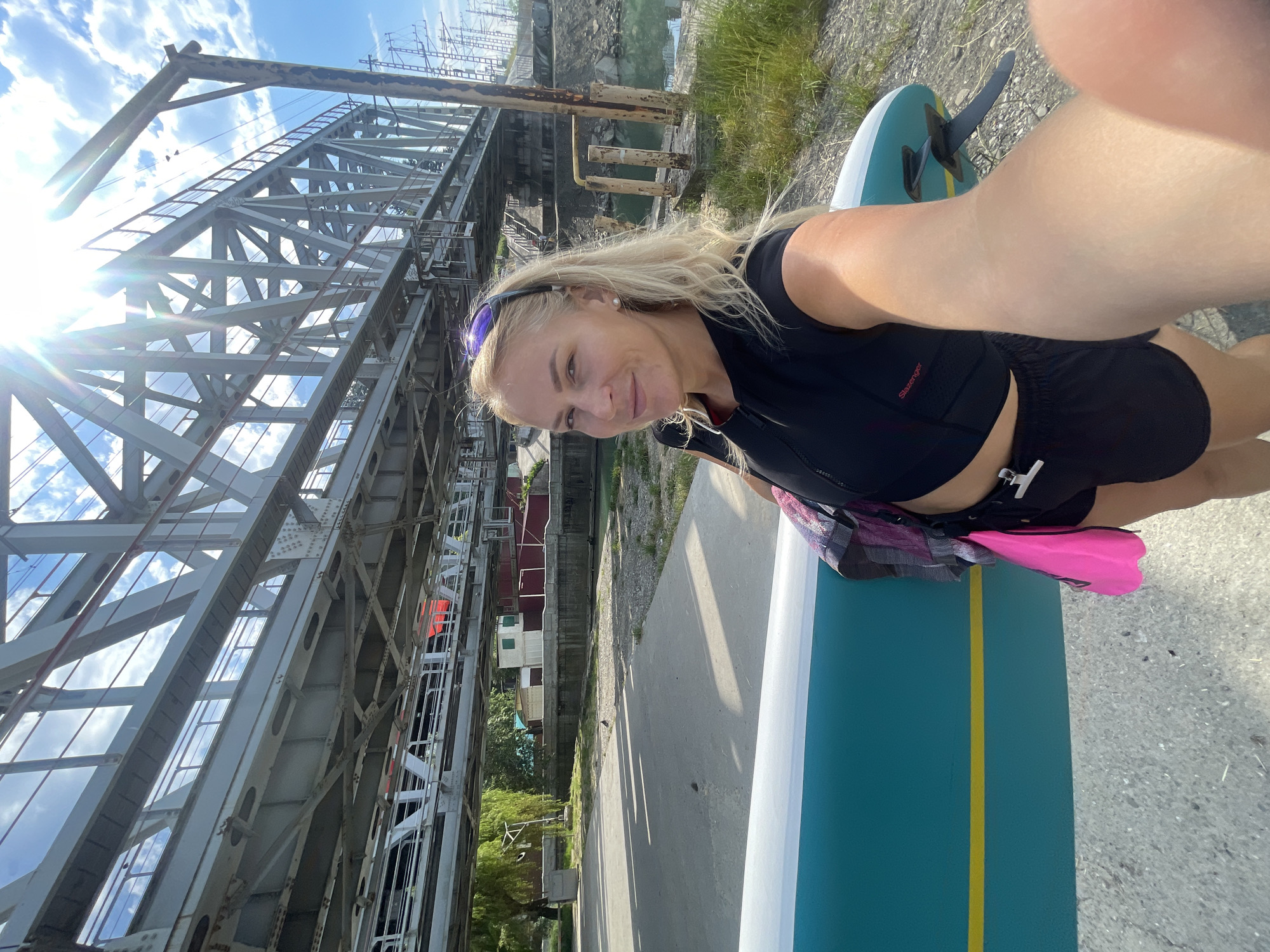 A woman with blonde hair and sunglasses on her head is taking a selfie outdoors. She is wearing a black crop top and black shorts, holding a teal and yellow stand-up paddleboard. Behind her is a large metal railway bridge. The environment appears to be near a body of water, with grassy and rocky terrain. The sky is blue with some clouds and the sun is shining brightly. Based on the presence of the railway bridge which spans the Mzimta River, this location is likely the Adler district of Sochi, Russia, possibly near the Mzimta River embankment.