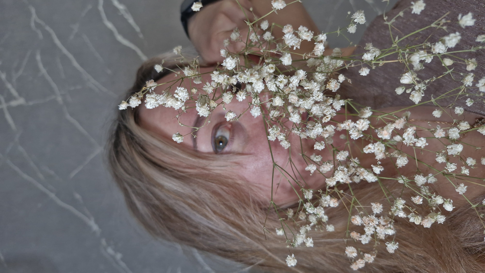 A person with blonde hair and green eyes is holding a bouquet of baby's breath flowers in front of their face. The background is a gray, textured surface resembling marble. A watch is visible on the person's wrist in the upper left corner.