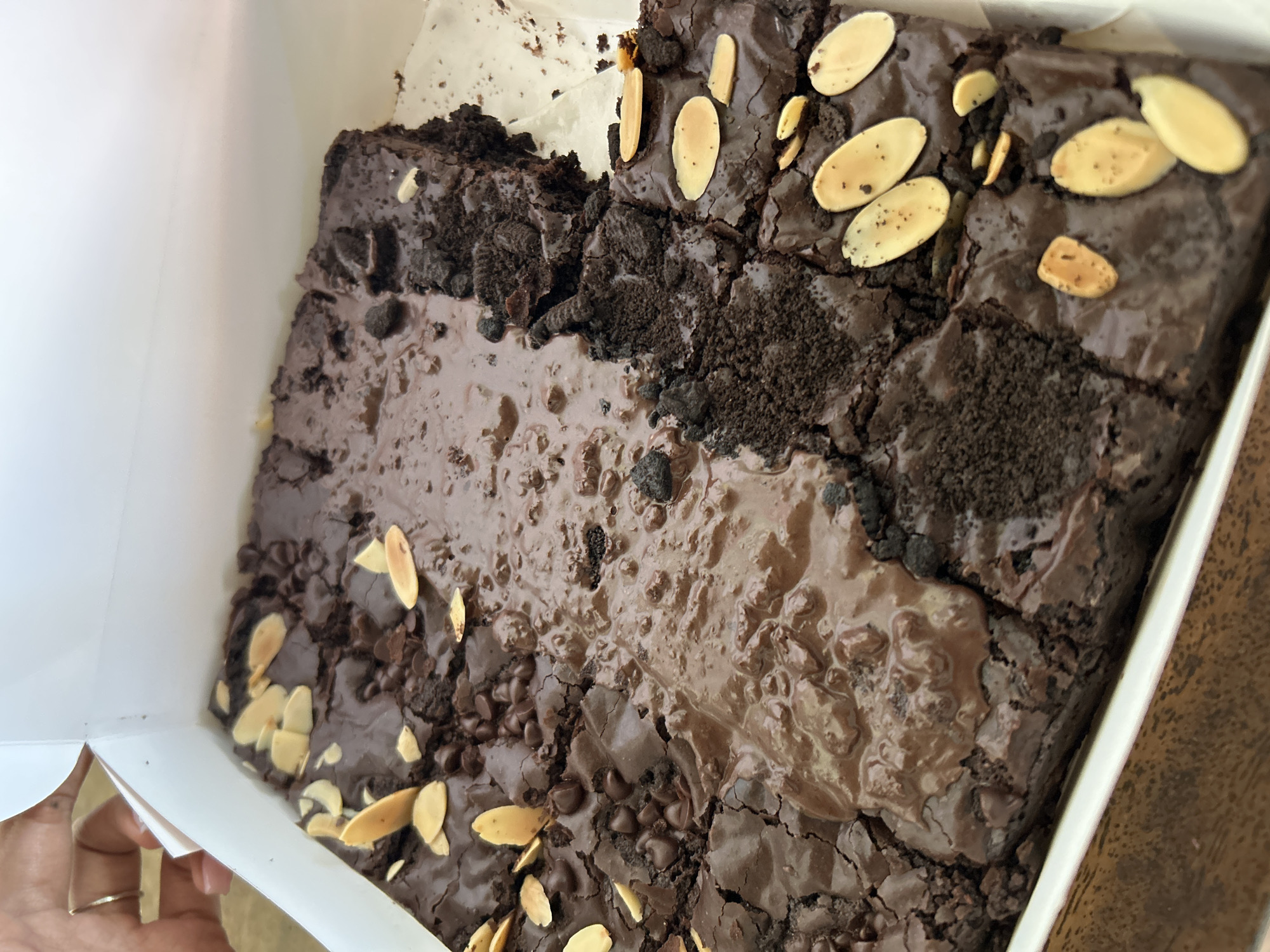 This media file shows a close-up, overhead view of a box of freshly baked brownies. The brownies are rich, dark chocolate, with a cracked surface suggesting a chewy texture. They are topped with a generous swirl of melted chocolate and a scattering of sliced almonds and mini chocolate chips. Some of the brownies also appear to have crushed cookie pieces mixed in, adding another layer of texture and flavor. The box is white and lined with white paper, typical for bakery packaging. The lighting suggests it is daytime, and the overall impression is one of delicious indulgence. The brownies are the sole focus of the image, with no people clearly visible, though a hand with a silver ring is partially visible on the left side of the frame, holding the edge of the box, suggesting someone is about to enjoy them. The background is blurred, but appears to be a wooden surface, possibly a table. There is no visible text in the image. The setting is likely a home or a bakery in Kuta, Indonesia, given the provided location context.