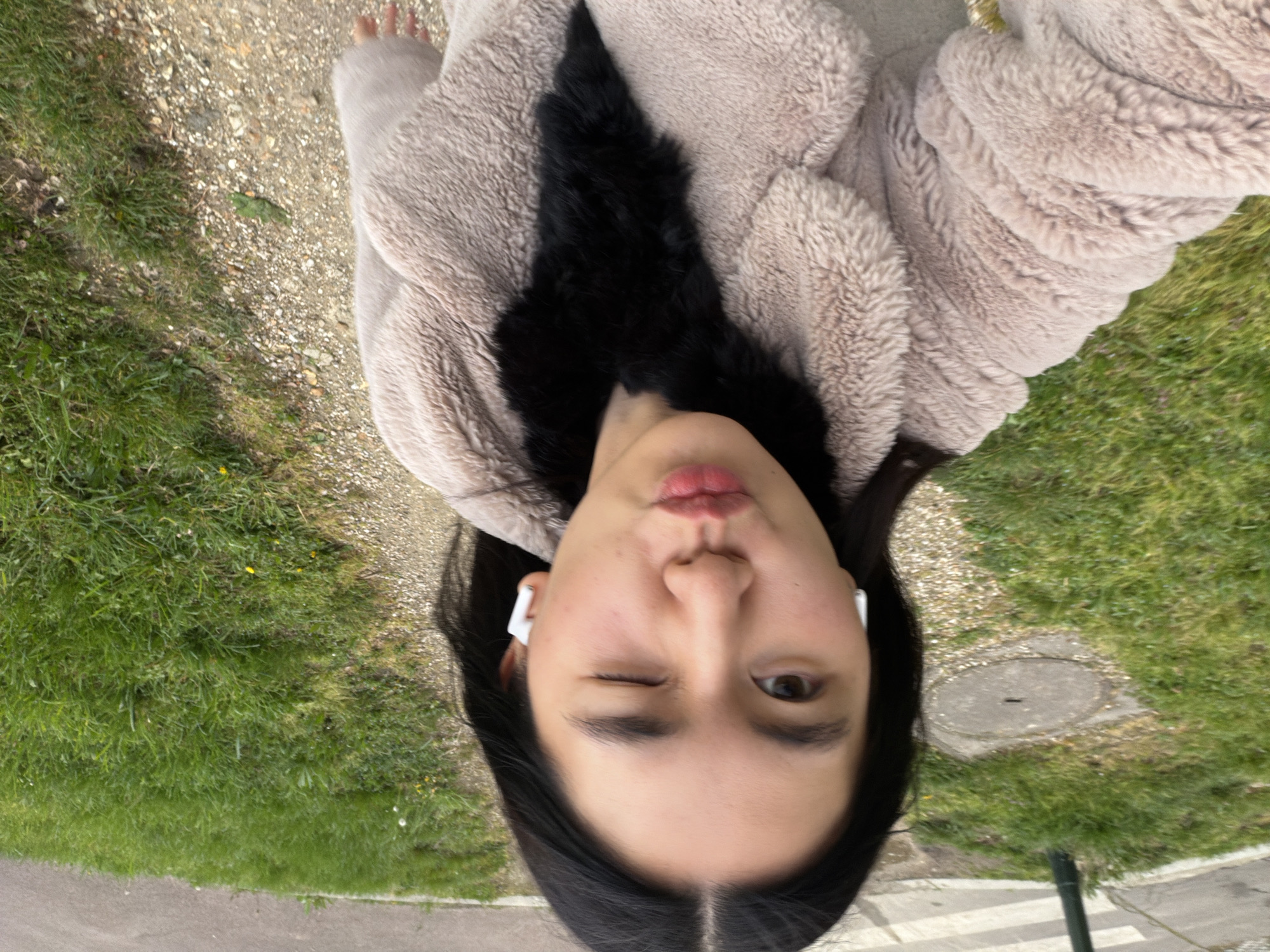 A woman stands outside wearing a light-colored furry jacket with a black fur trim. She is wearing white wireless earbuds and winking at the camera. The background shows a grassy area next to a gravel path and a paved road. There is a manhole cover visible in the grass. The location is likely in Paris, France.