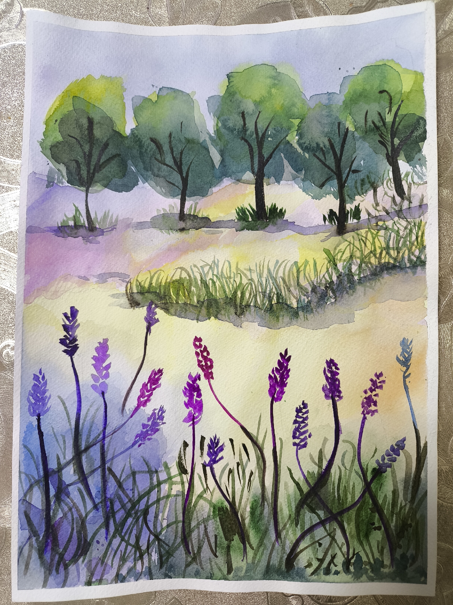 The image is a watercolor painting depicting a landscape with trees and lavender.
In the background, there is a row of five trees with green foliage and dark trunks. The trees are situated on a slightly elevated area with some grassy vegetation at their bases. Behind the trees, the sky is light blue.
In the foreground, lavender plants with purple flower spikes and dark green grass are visible. The grass appears to be watercolor paint with some blending of dark green, blue, and dark colors. The lower part of the painting is predominantly purple.
The painting style is loose and expressive, with visible brushstrokes and blending of colors. The overall atmosphere is bright and peaceful.
There is no indication that this is a photo taken in the city of Khimki, Russia.