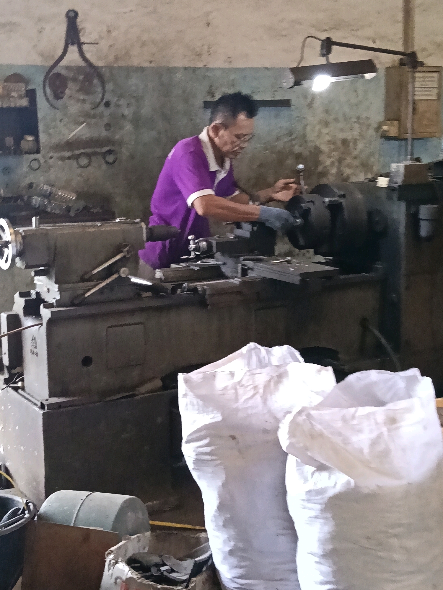 In a dimly lit workshop in Belimbing, Indonesia, a man wearing a purple collared shirt and glasses operates a metal lathe. The workshop appears cluttered, with tools and machinery covering the surfaces. A bright overhead lamp illuminates the immediate workspace. In the foreground, two large white sacks are visible, partially obscuring the lower portion of the scene. The walls are textured and appear aged, with various items, including a caliper-like tool, hanging on them.