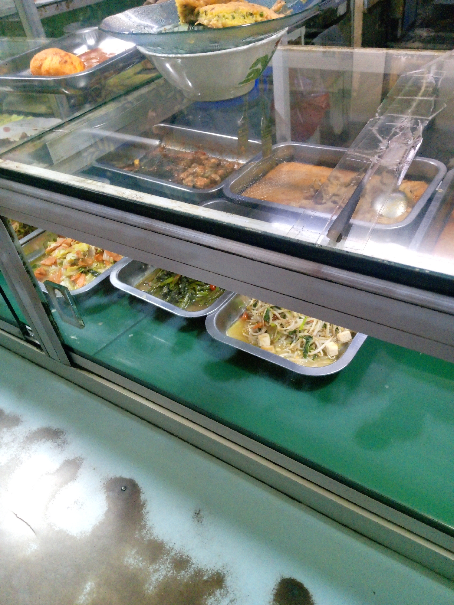 The image shows a food display case filled with various dishes, likely in a food stall or restaurant in Bitung Jaya, Indonesia. The display case has a glass front and is divided into compartments holding different food items in metal trays.
In the top left, there are two round, golden-brown items, possibly fried eggs or meatballs, in a rectangular tray. To the right of this, a large bowl contains a yellowish food item, possibly fried rice or a dessert.
Below the top section, several more trays are visible. One tray contains a dish with meat and sauce. Another tray holds what appears to be stir-fried vegetables with carrots and cabbage. A further tray contains a stir-fry of bean sprouts and cubes of tofu. A tray to the right appears to contain a dark, saucy dish, possibly meat.
The display case is illuminated from above, casting light on the food. The background is blurred but suggests an indoor setting with some shelving and possibly other food items. The overall impression is of a busy food service area, common in market settings in Indonesia.