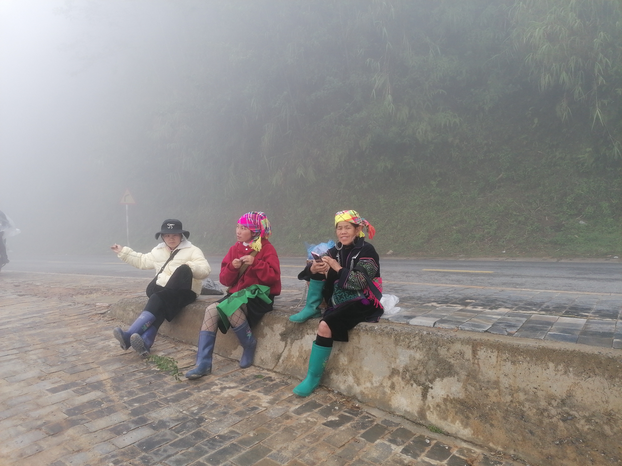 Three women are sitting on a low wall that runs along a roadside. The woman on the left wears a black hat, white jacket, black skirt, and blue boots. She is holding something in her right hand. The woman in the middle is wearing a pink and white patterned headscarf, a red sweater, a black skirt, green stockings, and blue boots. She is holding something in her hands. The woman on the right wears a yellow and red patterned headscarf, a black jacket with colorful embroidery, a black skirt, and green boots. She is holding a cell phone. The background is foggy, with green vegetation visible. The location is Unknown, Unknown.