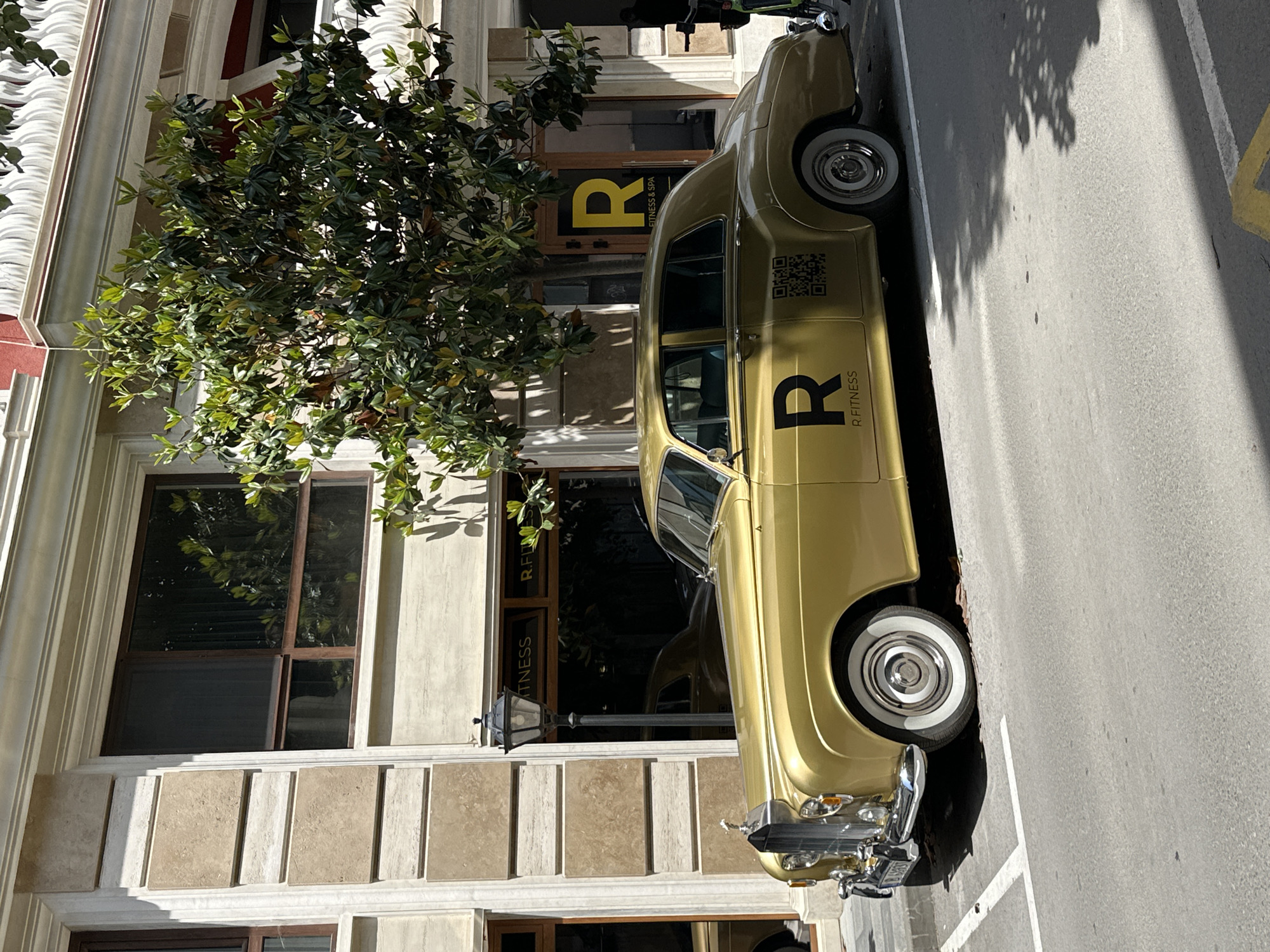 The scene shows a vintage, gold-colored car parked on a street next to a building in Varna, Bulgaria. The building has a sign that reads "R Fitness". The car's body has a large "R" logo, and a QR code. The building is light-colored, with visible architectural details like windows and columns. A tree obscures part of the building's facade. The street surface is asphalt, and a shadow is cast on the road.