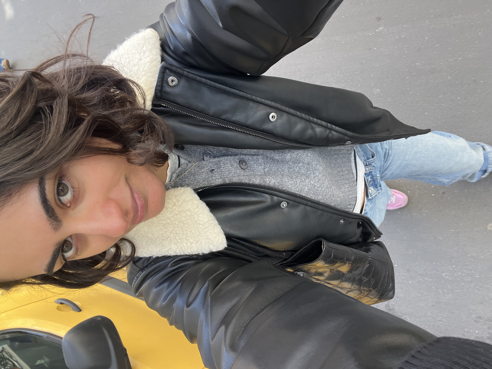 A woman with dark hair is taking a selfie outdoors. She is wearing a black leather jacket with a white shearling collar over a gray sweater and light blue jeans. She has a black handbag on her shoulder and is standing next to a yellow car, likely a taxi. In the background, there is a gray street. The presence of the yellow car, the style of dress, and the general surroundings suggest that the location is likely Paris, France.