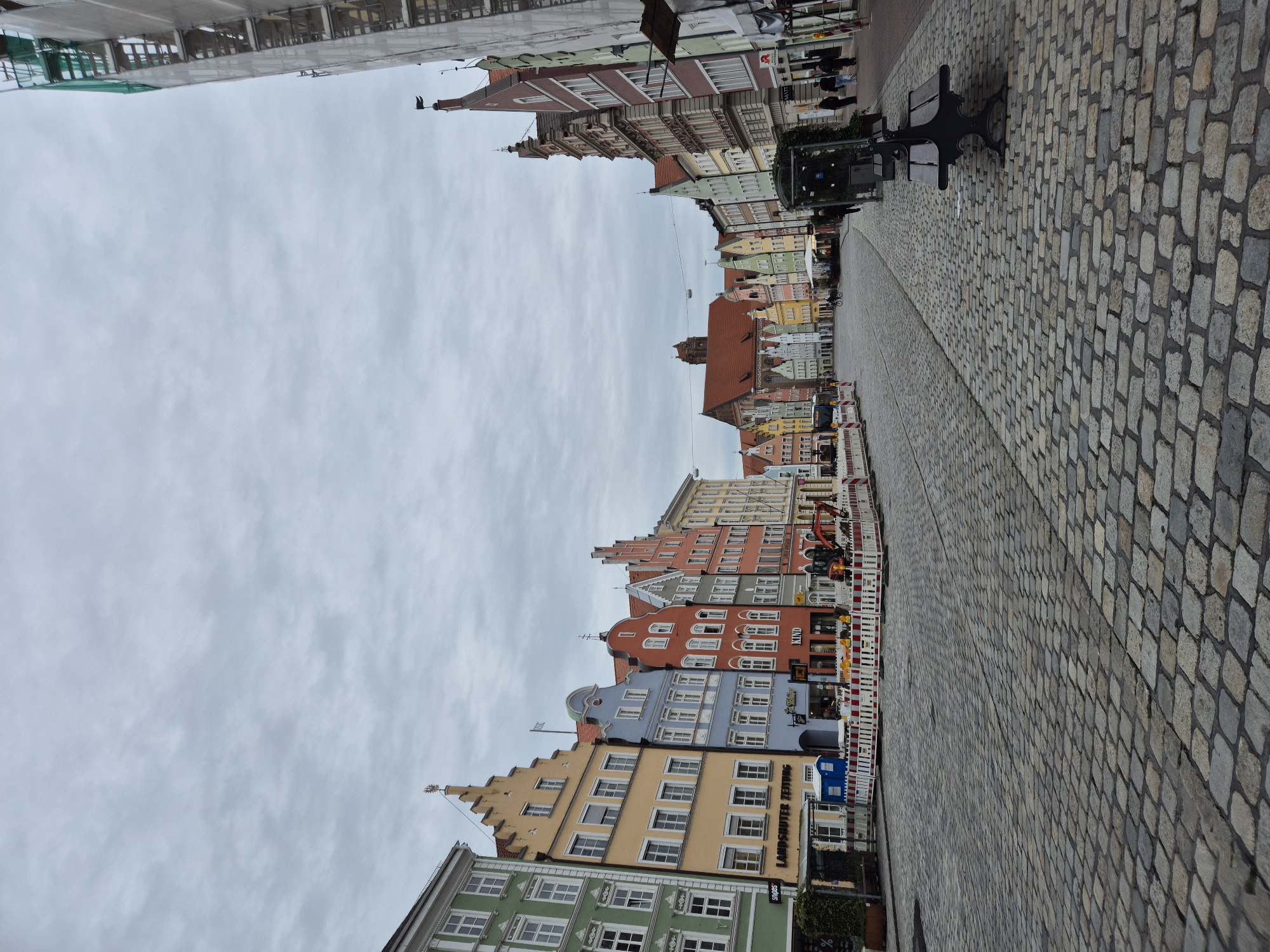 This image depicts a street scene in Landshut, Germany, likely the historic city center, characterized by cobblestone paving and colorful, gabled buildings. The sky is overcast, contributing to a subdued lighting condition. On the left side of the street, a row of vibrantly colored buildings stands with traditional architecture, featuring multi-story facades and distinct rooflines. Signs for "SNOPPA," "LANDSHUTER ZEITUNG," and "KIND" are visible, indicating retail or commercial establishments. Construction barriers with red and white stripes are present in front of some buildings, suggesting ongoing work or renovation, and an excavator is visible behind the barriers. On the right side of the street, the perspective leads towards more buildings, including one partially covered in scaffolding and white sheeting, indicating construction or maintenance. A dark metal park bench is situated on the right side of the cobblestone street, with a glass-enclosed structure beside it. Several people are visible in the distance, walking along the street or near the buildings. A bicycle is also seen in the middle of the street. The overall impression is of a European city square or main thoroughfare on a cloudy day, with elements of both historic charm and ongoing urban development.