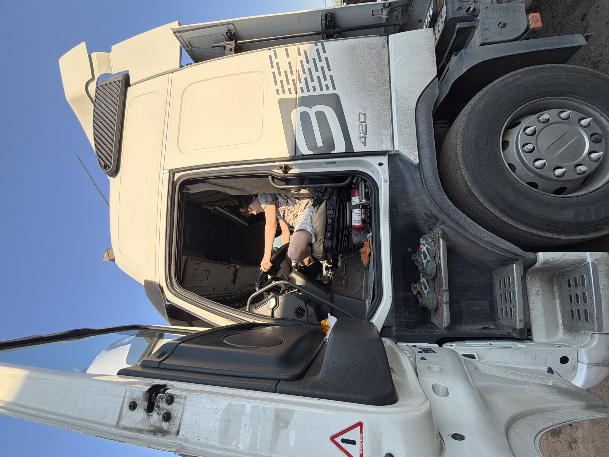 The image shows a person seated inside the cabin of a white Volvo 420 truck. The truck's passenger door is open. The background suggests an outdoor environment with a clear, blue sky. The location in the city of Chaikovsky, Russia, is not discernible from the visual information provided.
